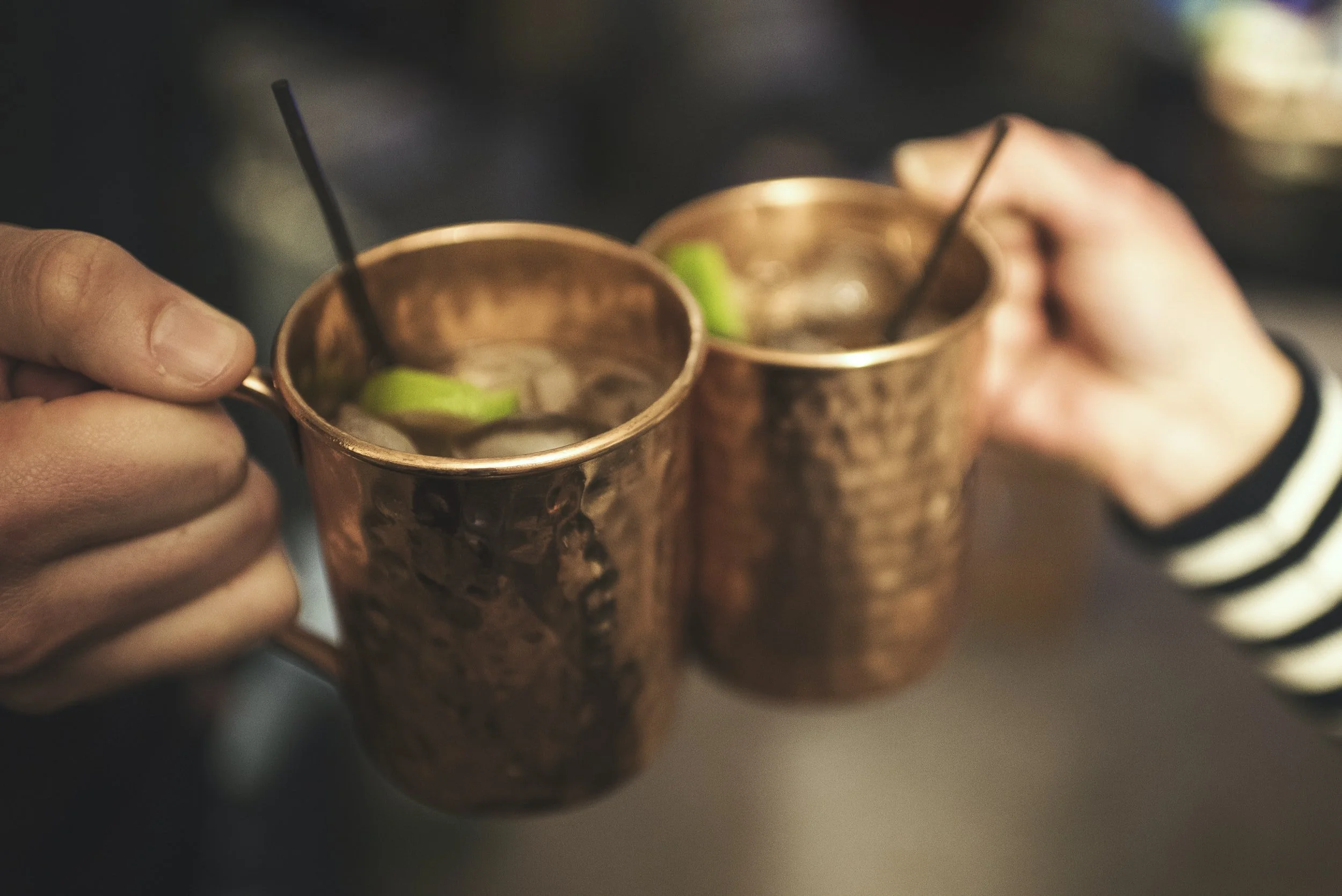 Two hands holding copper mugs with lime slices and black straws, clinking together in a toast.