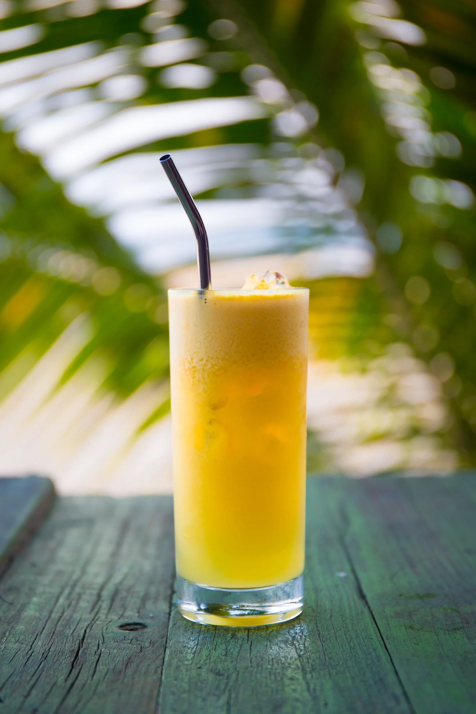 Tall glass of yellow mango smoothie with a black straw, placed on a weathered green wooden surface, with tropical green leaves in the blurred background.