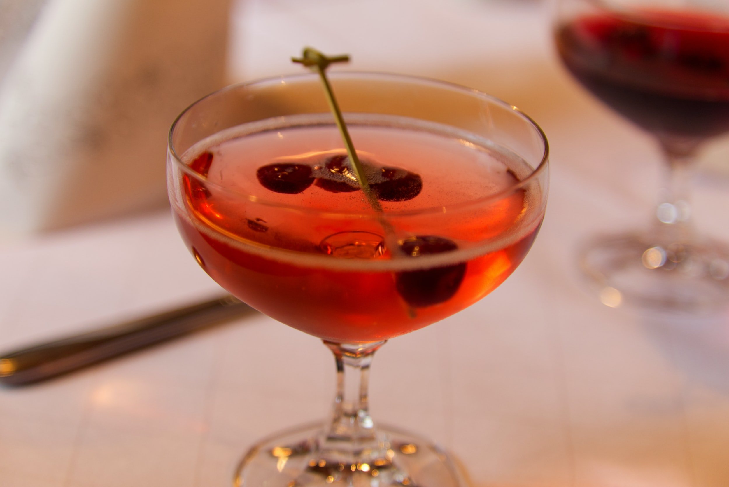 A cocktail glass of red-colored drink garnished with black cherries and a green cherry stem, on a white table.