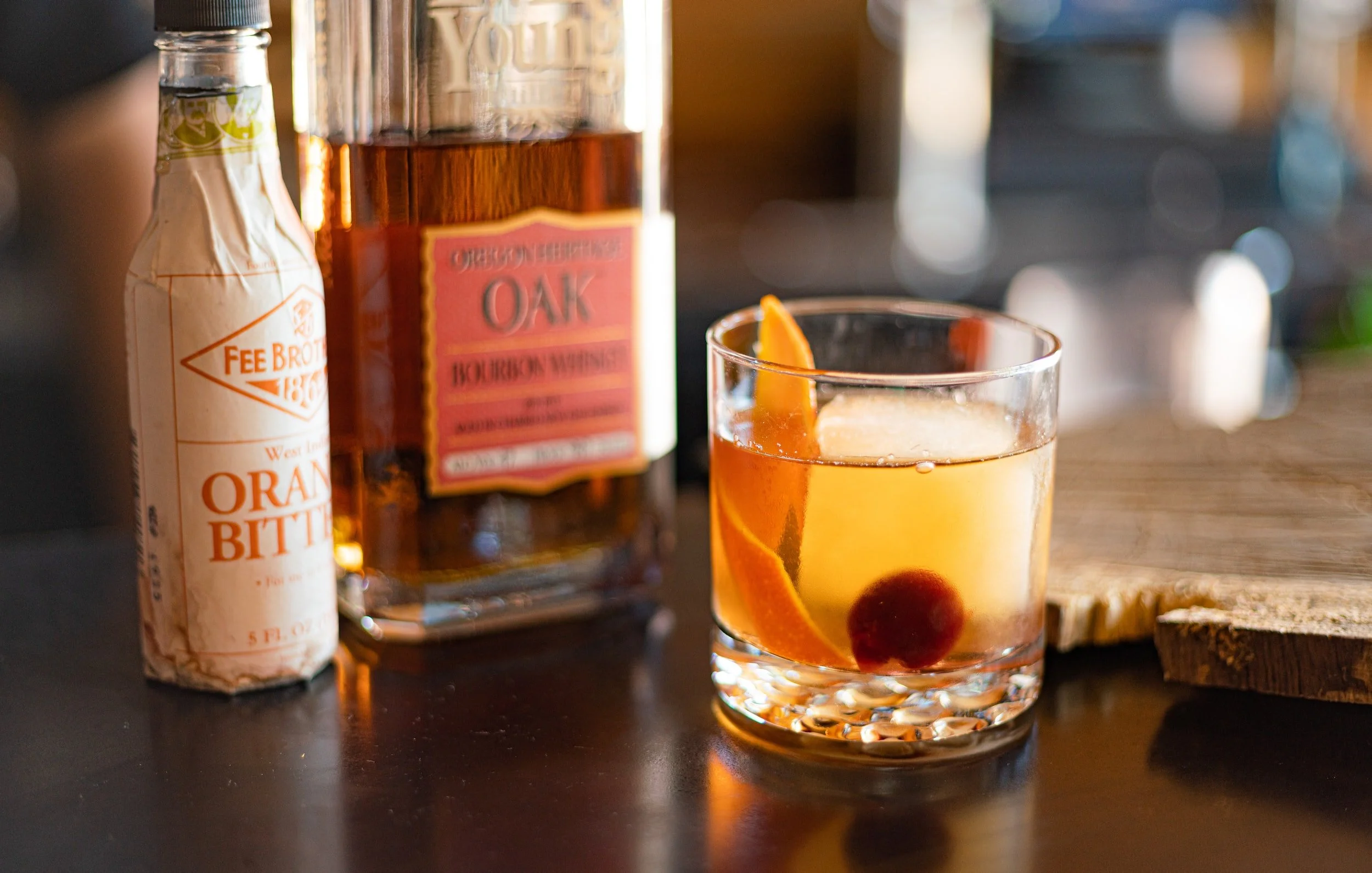 A glass of cocktail with an orange slice and cherry, with a bottle of bourbon whiskey and a bottle of orange bitters nearby on a dark table.