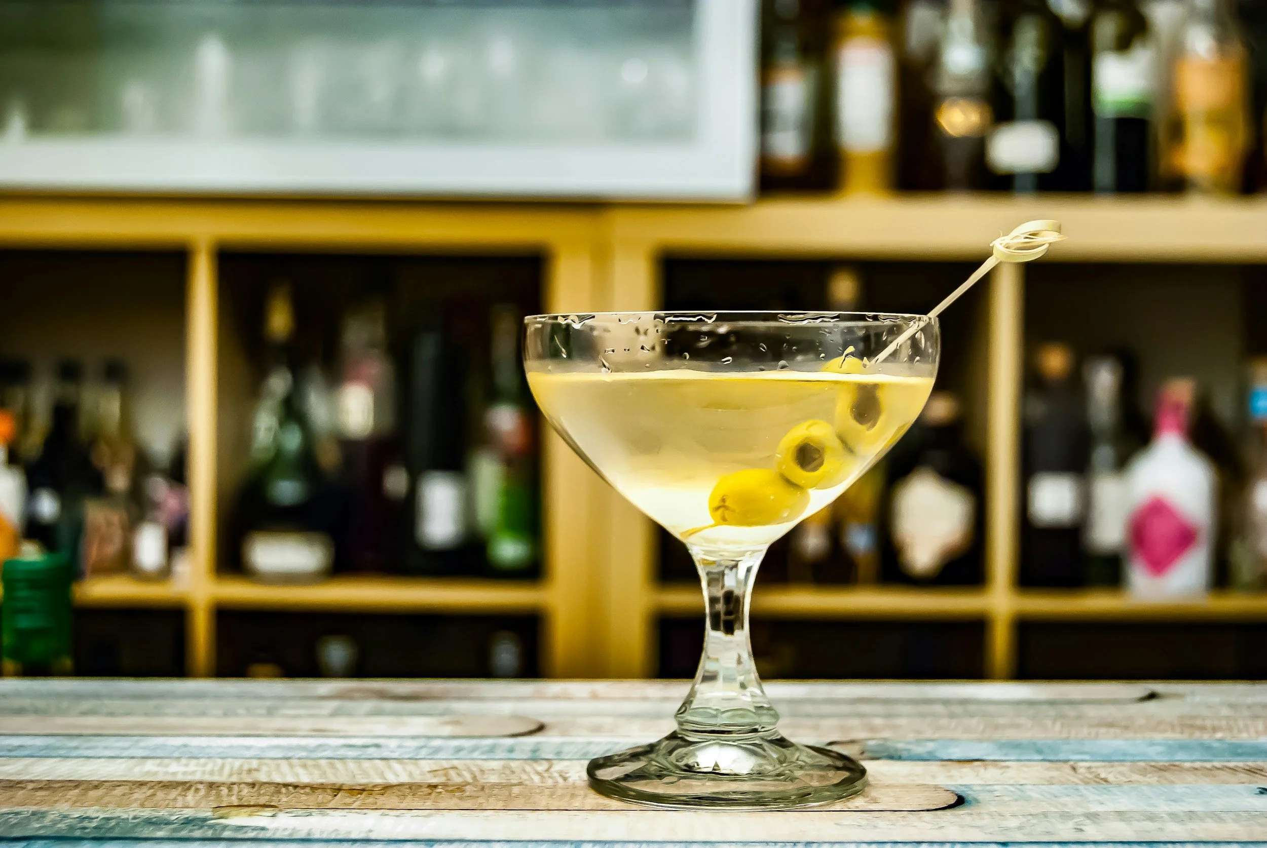 A martini glass with green olives and a cocktail pick on a wooden bar counter, blurred liquor bottles in the background.