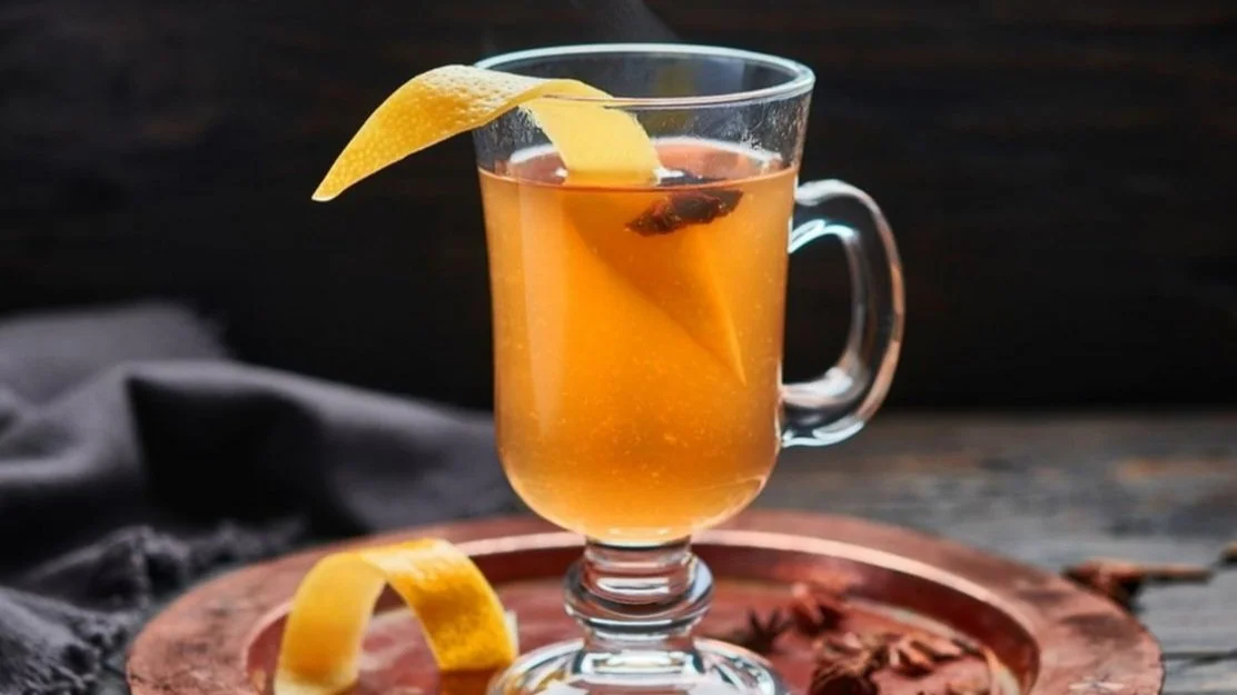 A clear glass mug filled with amber-colored hot tea garnished with a lemon wedge, set on a round copper tray with additional lemon slices and cinnamon sticks on a rustic wooden surface.