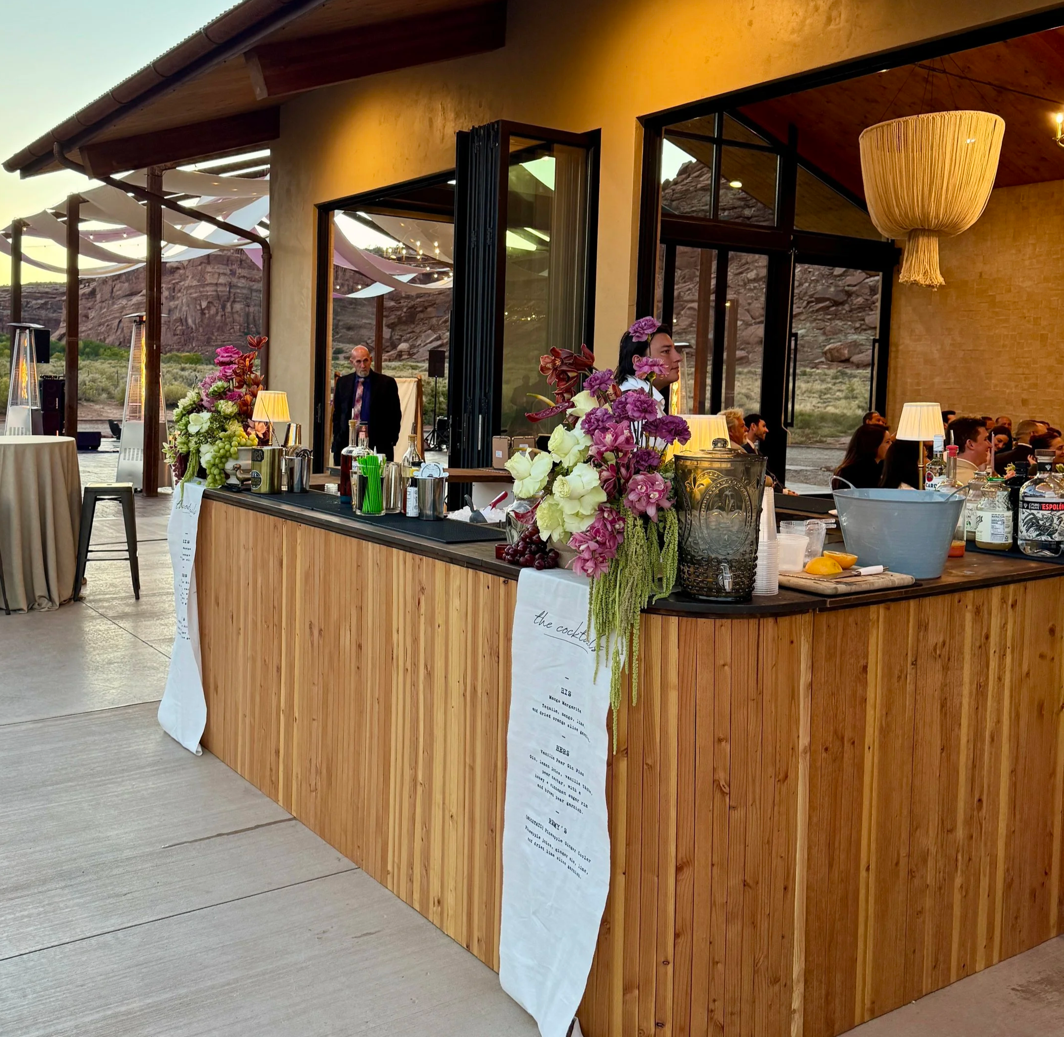 Outdoor wedding reception bar with pink and white flowers, alcohol bottles, and a menu, set against a desert landscape at sunset.