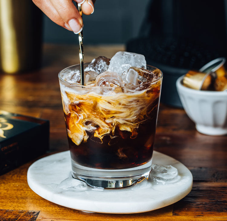 A glass of iced coffee with cream swirling in it on a marble coaster, with ice cubes on top and a hand stirring with a silver spoon.