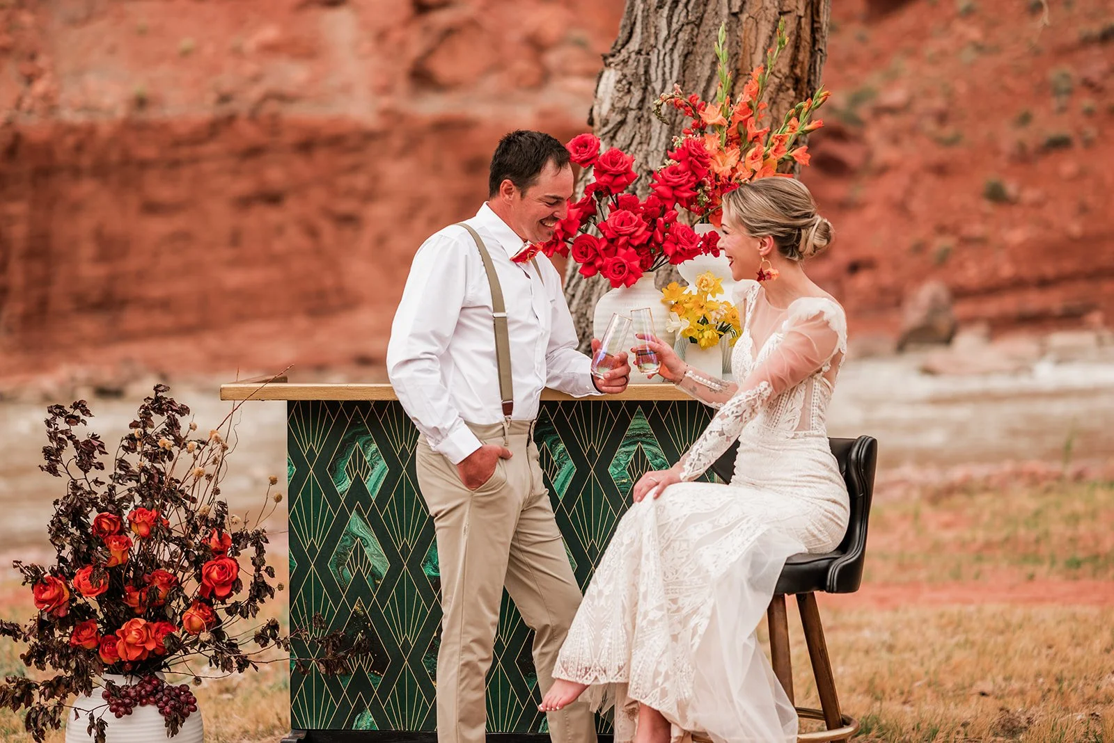 A wedding couple sharing a toast with glasses of champagne outdoors, with a desert landscape background and floral arrangements.