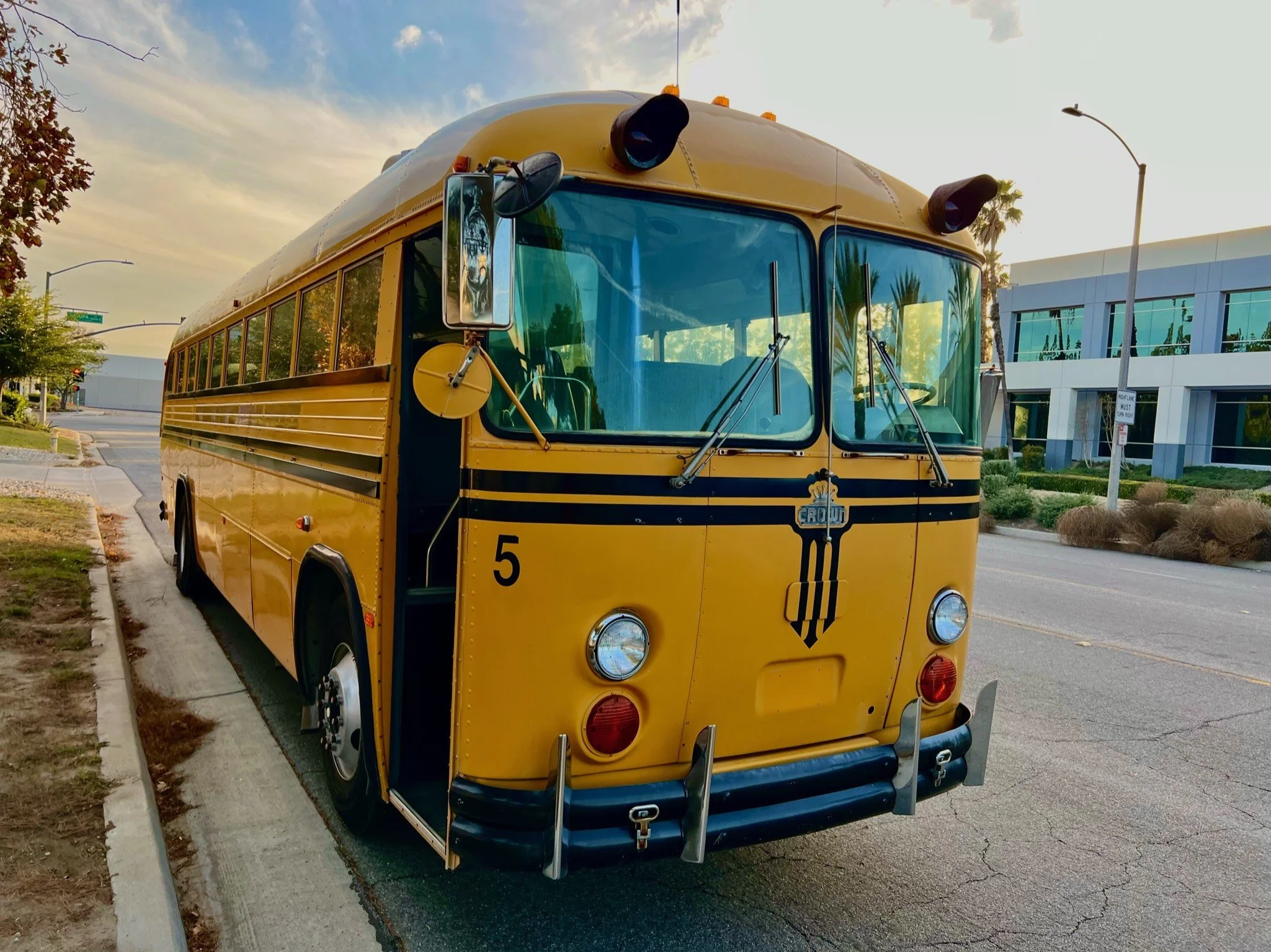Yellow vintage school bus parked on the side of the road with a modern building and trees in the background.