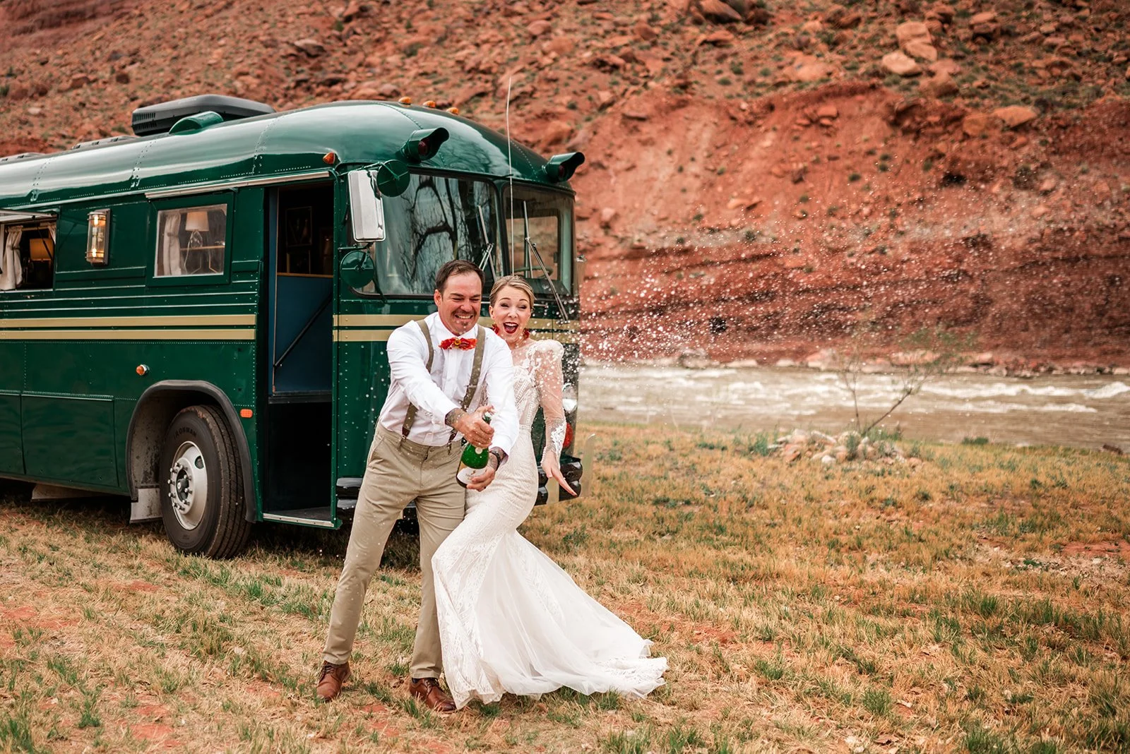 A newlywed couple celebrating outdoors near a vintage green bus and a river with red rocky hills in the background. The groom and bride are smiling and holding a champagne bottle as they celebrate.