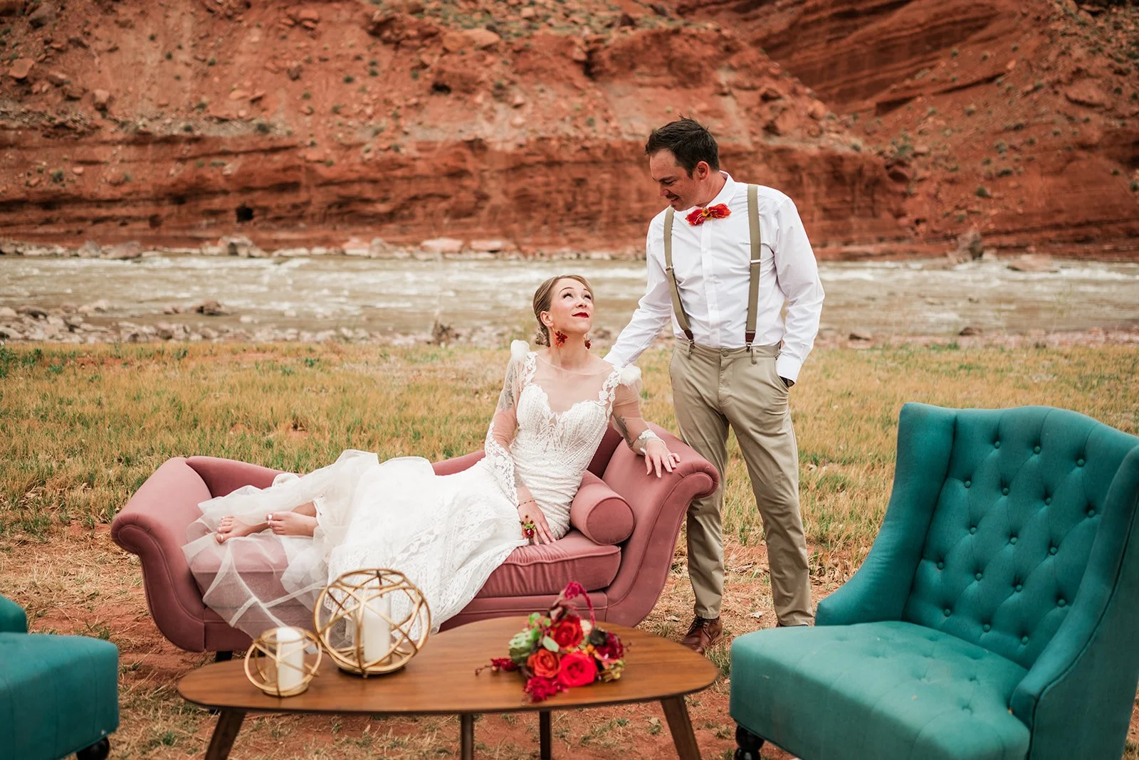 A woman in a wedding dress sitting on a vintage pink sofa, and a man in suspenders and khaki pants standing beside her, outdoor setting with rocky red cliffs in the background, a wooden table with flowers and candles in front of the sofa.
