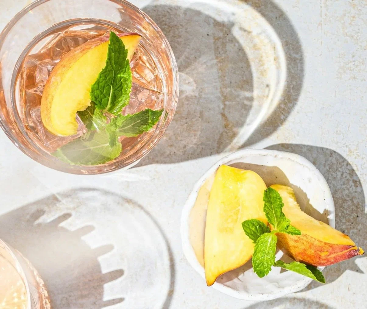 Overhead view of four glasses of pink lemonade with mint and lemon slices, a white plate with lemon wedges and mint, and two wooden drink stirrers on a light surface with shadows.