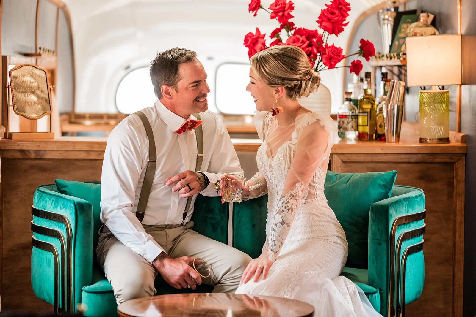 A couple dressed in wedding attire sitting on a green velvet couch inside a cozy room, smiling and looking at each other, with a woman holding a glass. The room has wooden furniture, red flowers, and a window in the background.