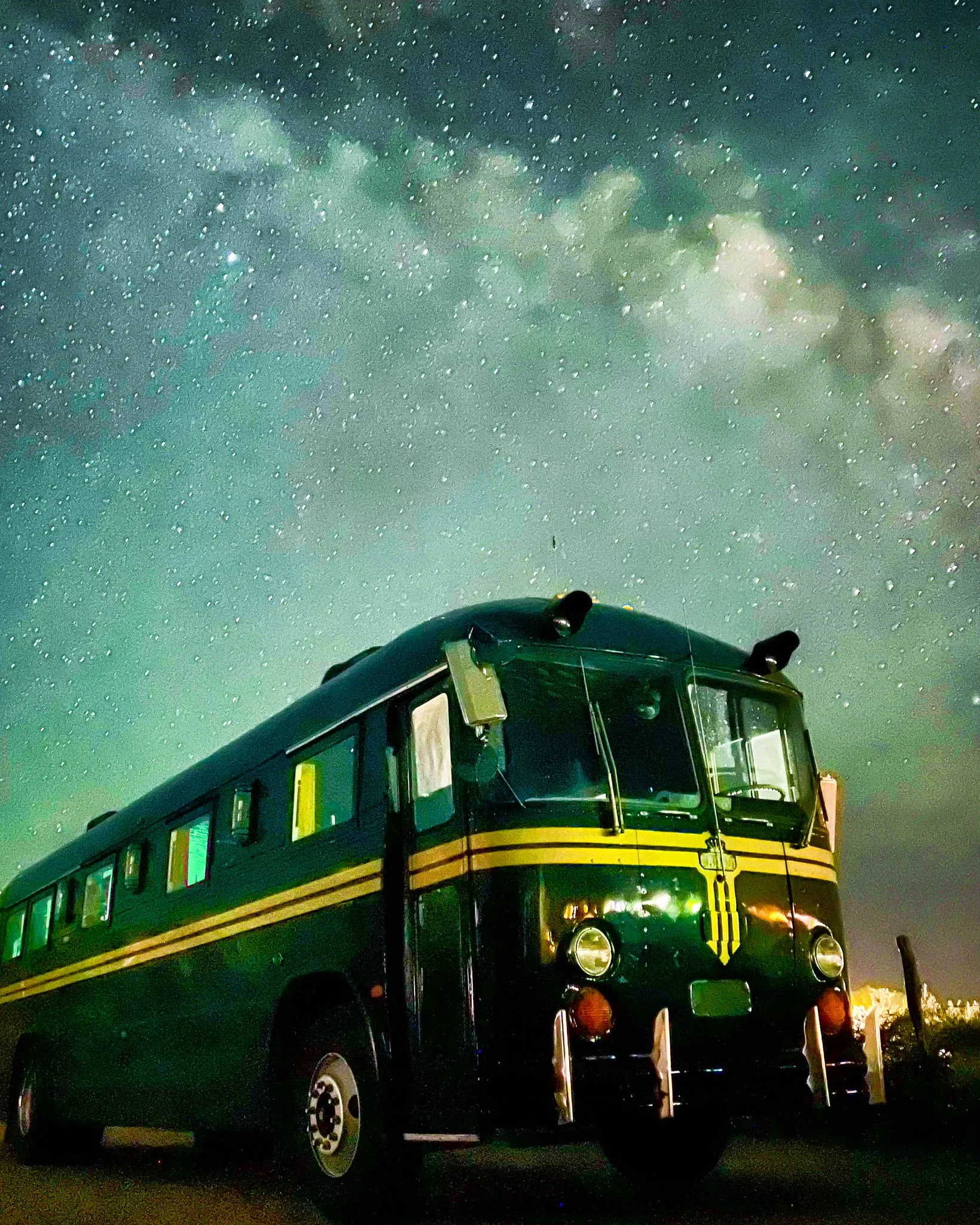 A vintage green bus with yellow stripes parked under a starry night sky with the Milky Way visible.