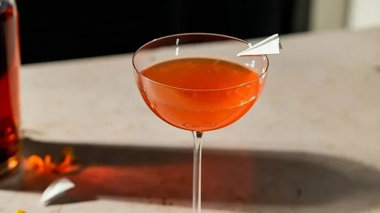 A pink or orange cocktail in a coupe glass with a small paper umbrella garnish.
