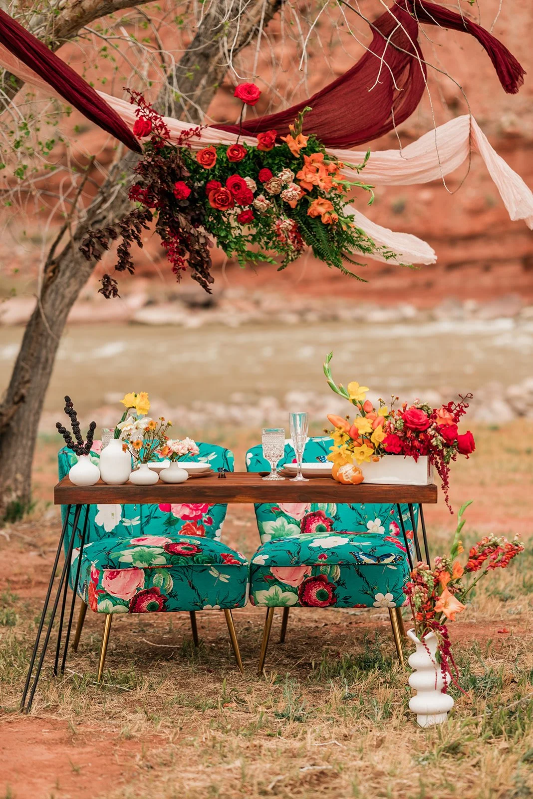 A decorated outdoor table setup with a wooden table and two floral patterned chairs, set against a natural background with stream and rocks. The table features vases with flowers and glassware, and above it, a canopy of fabric and flowers is suspende