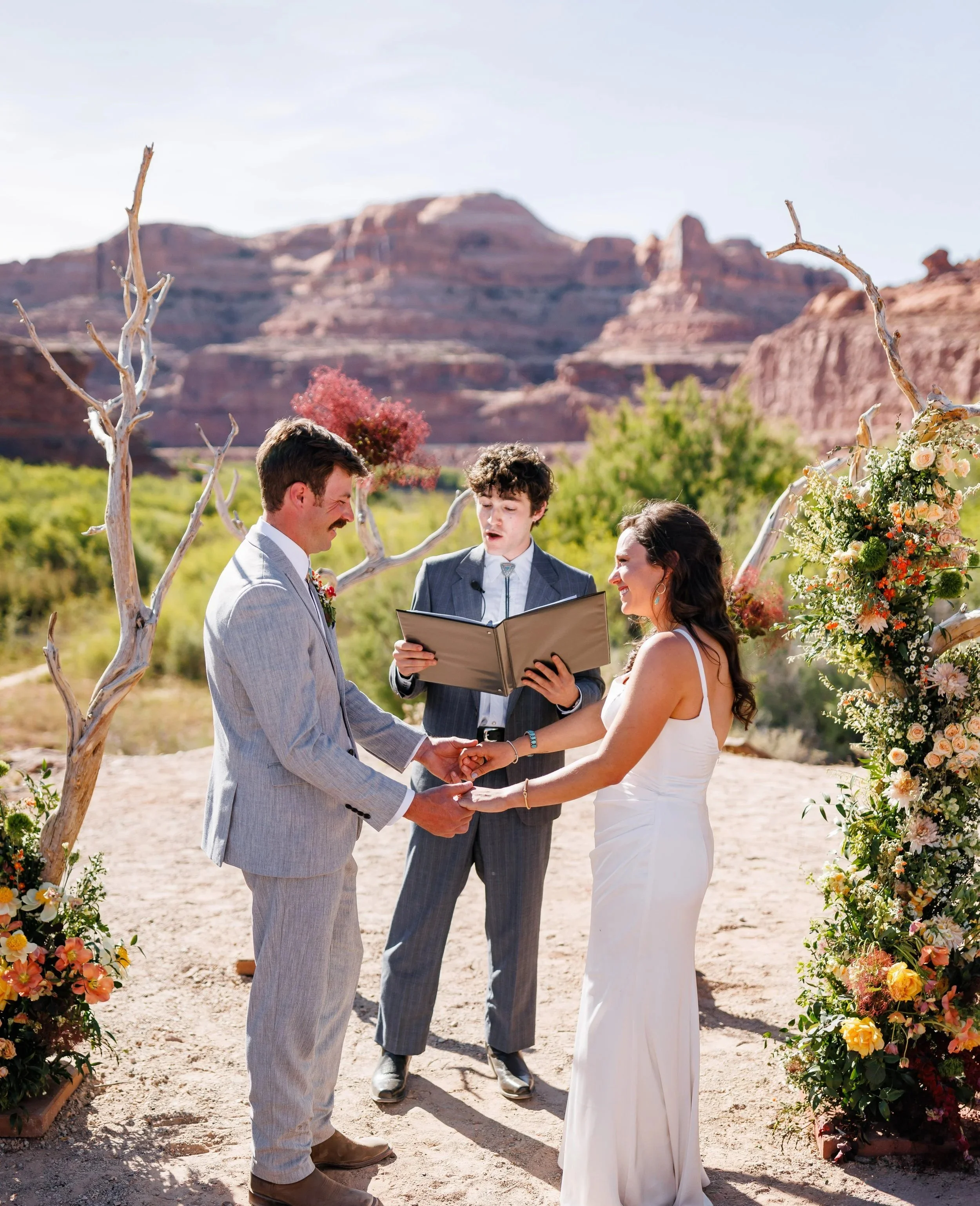 A couple getting married outdoors in a desert landscape, holding hands and exchanging vows. The officiant stands behind them, and there are flower arrangements and bare tree branches decorating the ceremony area. The background features mountains and a clear sky.