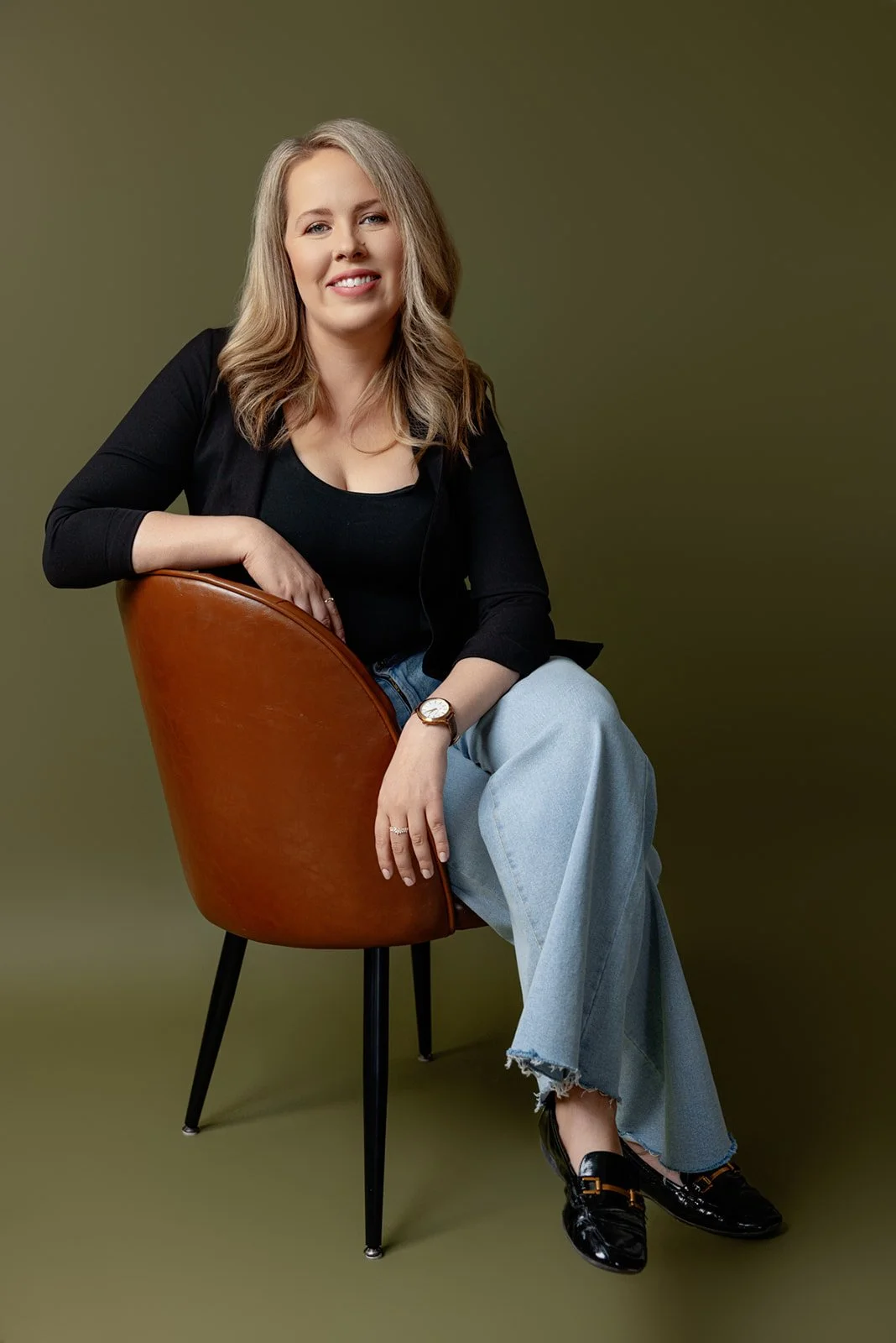 A woman with blonde hair sitting on a modern chair with wood and black legs, against a plain olive green background. She is wearing a black top, black jacket, light blue ripped jeans, black shoes, and a gold watch, smiling at the camera.
