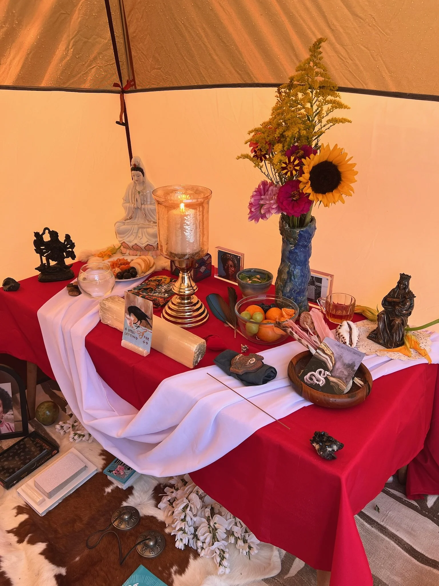 A multi-colored altar with religious and decorative items, including a ceramic Buddha, a sunflower, a candle, bowls of fruit, small statues, photographs, and flowers, on a red and white cloth inside a tent.
