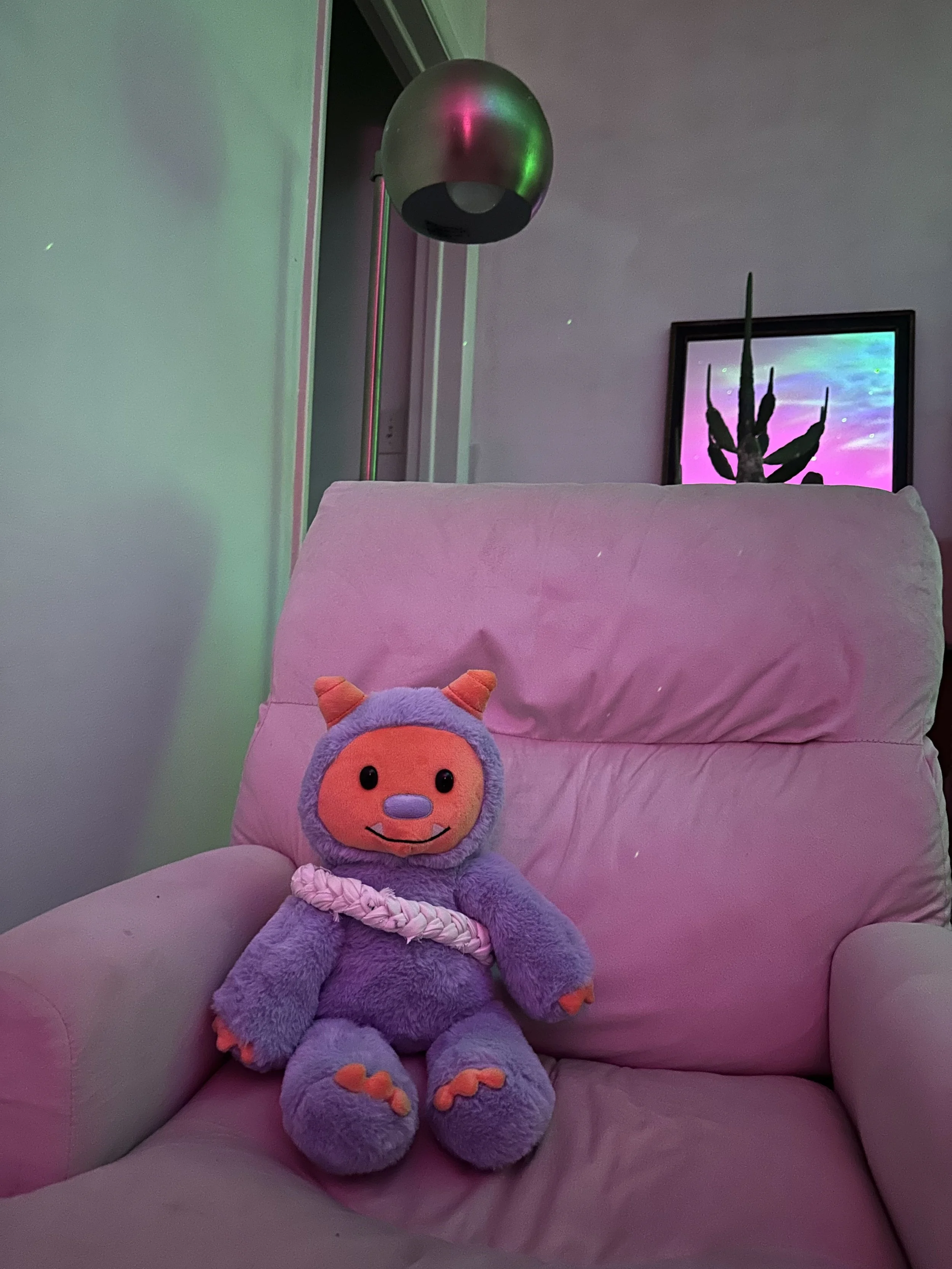A bear-like plushie with orange horns, orange hands and feet, and a smiling face sitting on a pink armchair. There is a framed picture of a cactus and a colorful sky on the wall behind. There is a grief braid wrapped around the plushie's torso.