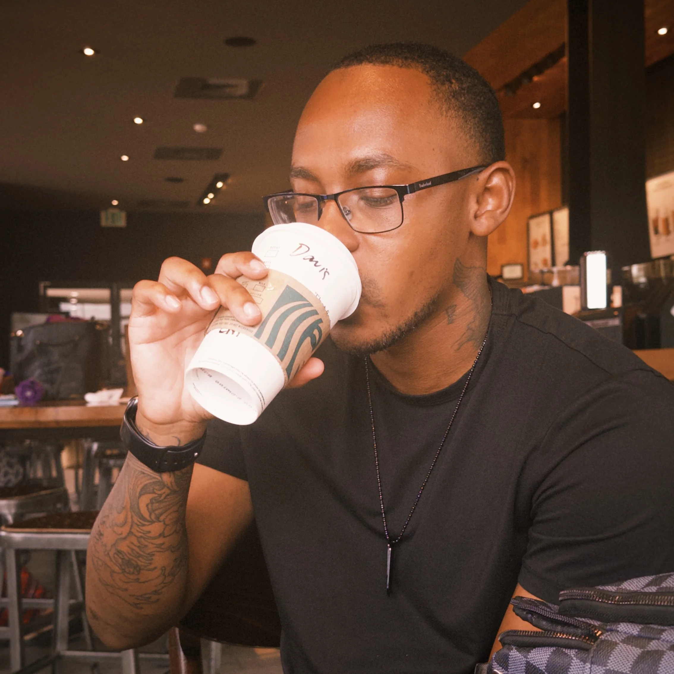 A man wearing glasses and a black t-shirt is drinking from a paper coffee cup in a cafe.