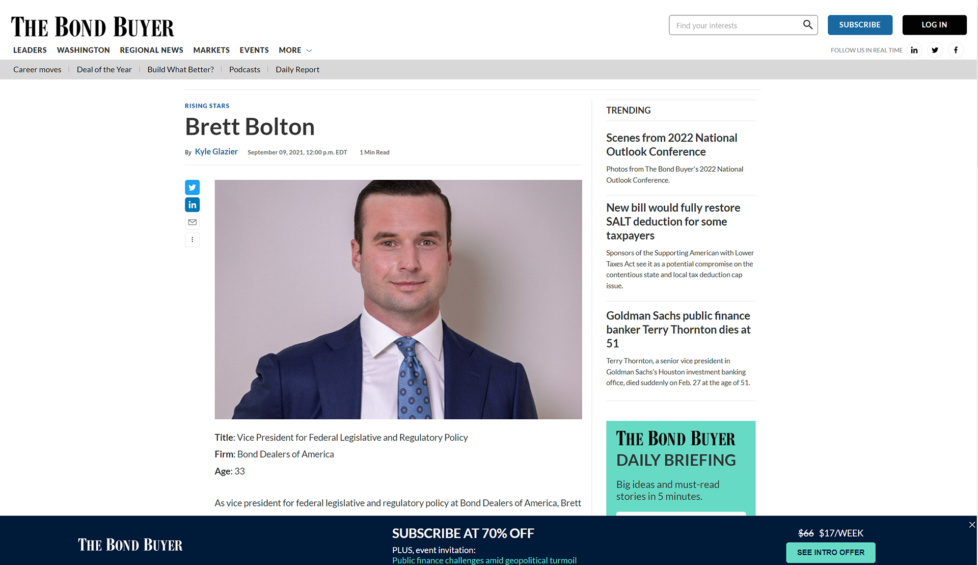 Professional headshot of Brett Bolton, a young man with short dark hair, clean-shaven, wearing a navy blue suit, white shirt, and a light blue patterned tie, smiling against a plain grey background.