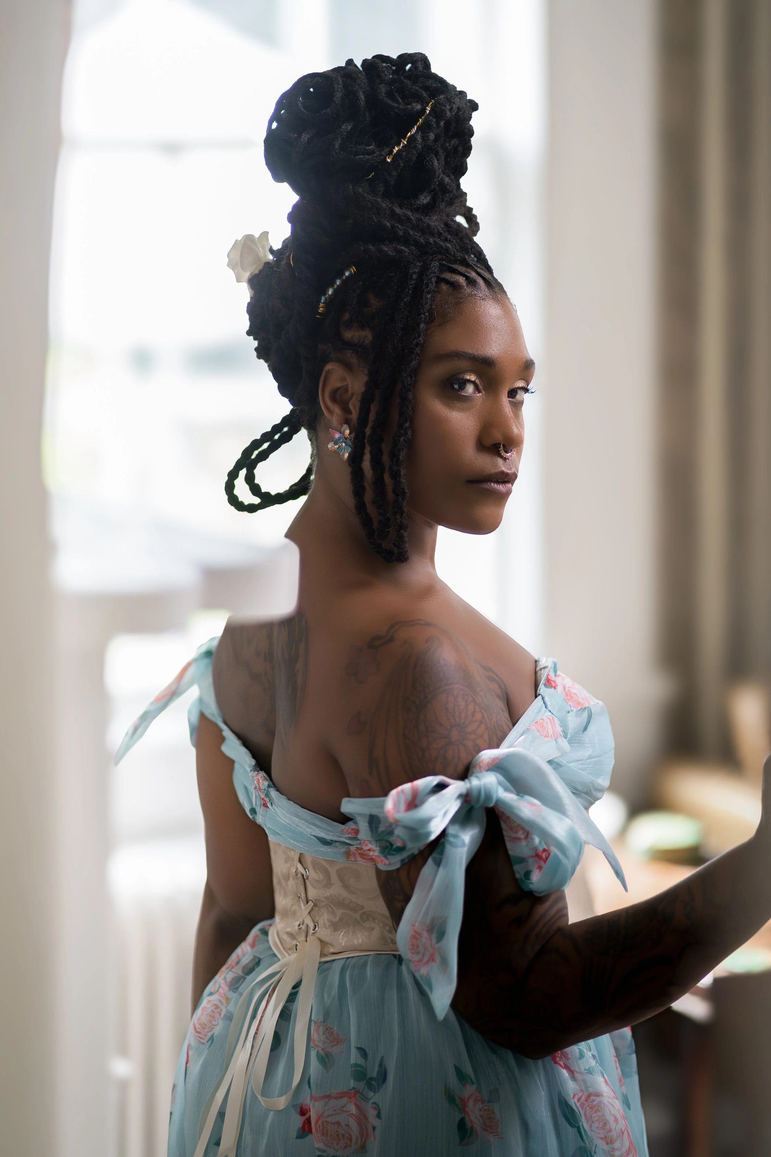 Woman in a floral blue dress with braided updo and tattoos, looking over her shoulder.