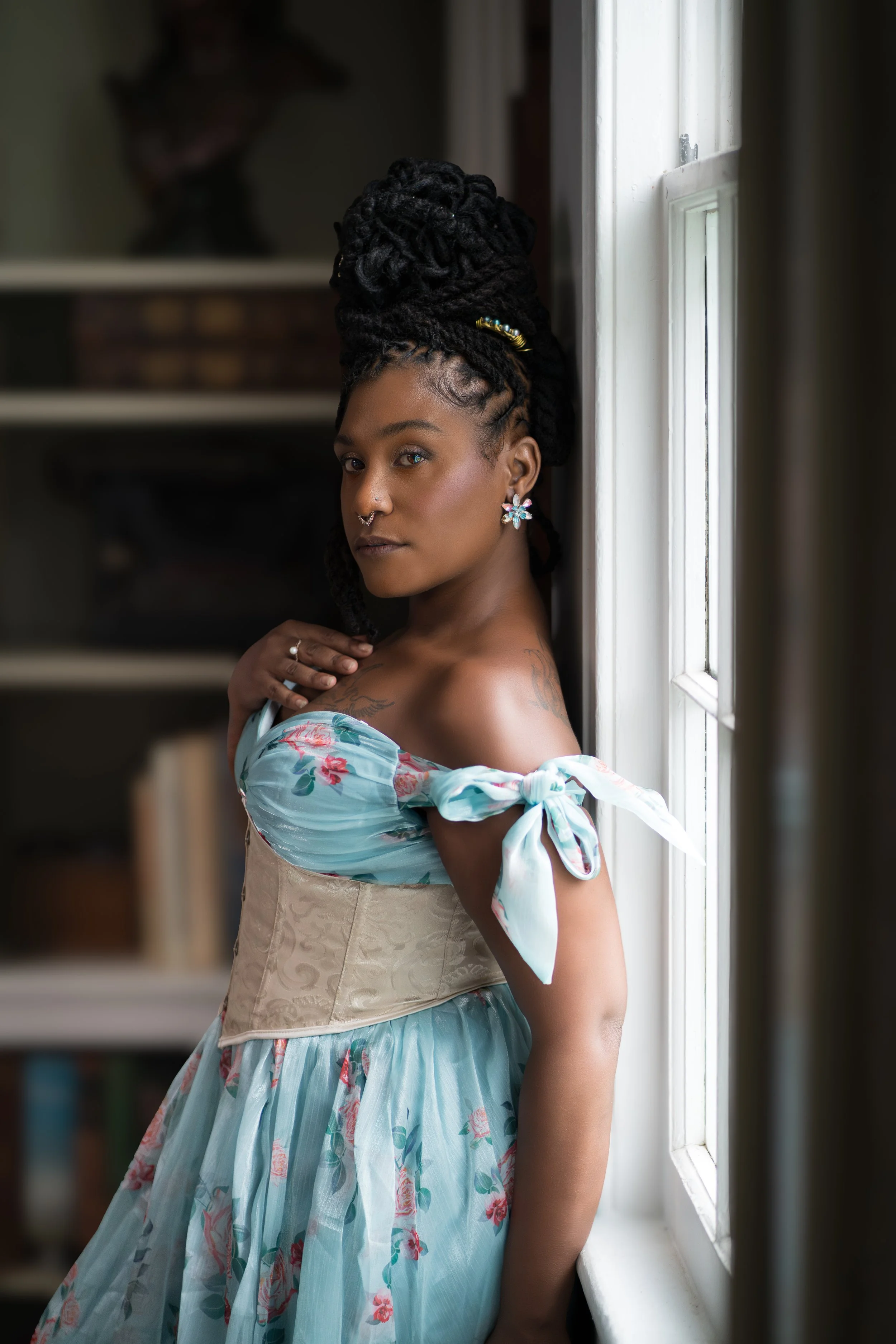 Person wearing a floral off-shoulder dress and corset, standing by a window.