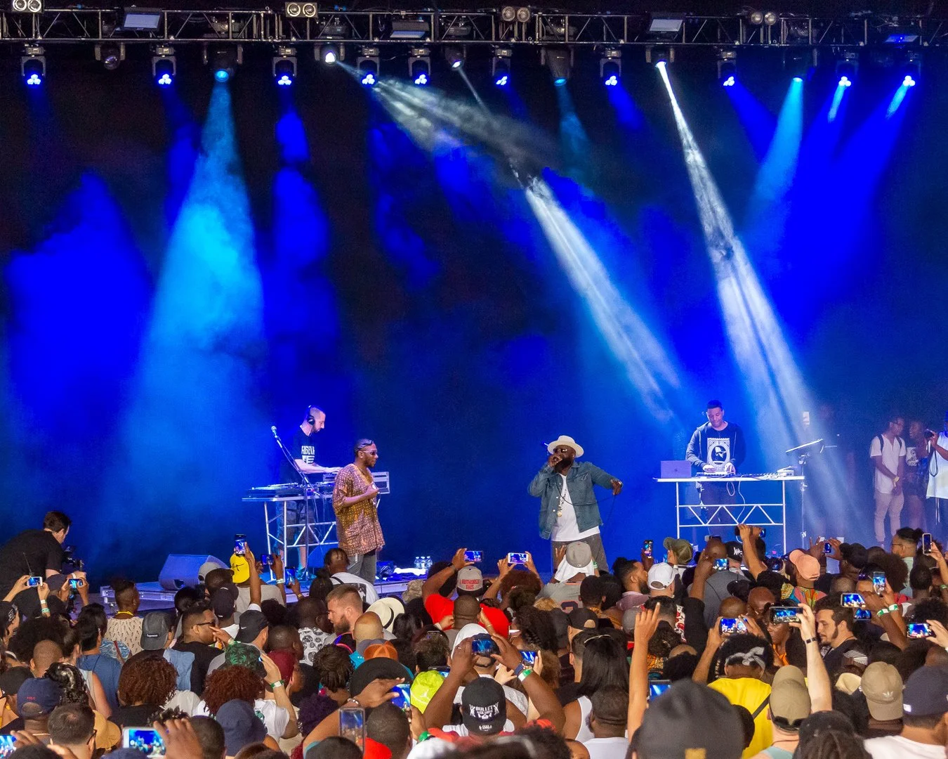 A concert on stage with musicians and a large crowd in front, many audience members holding up phones to record, stage lit with blue and white lights.