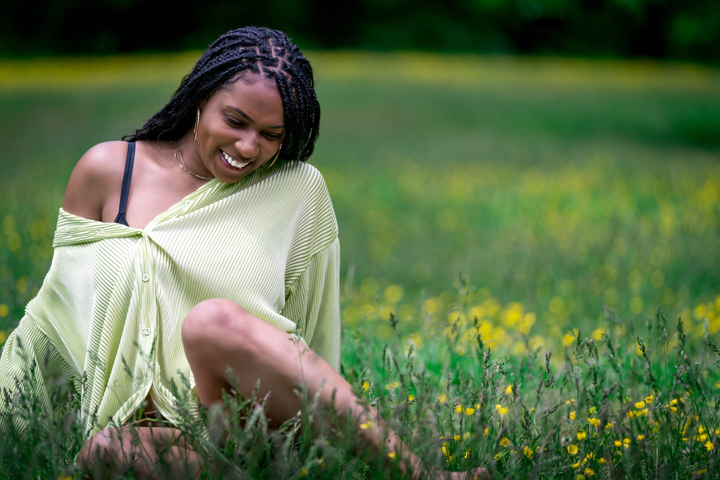 A woman with dark braided hair wearing a yellow off-shoulder shirt and black bra strap, sitting in a grassy field with yellow flowers, smiling and looking down.