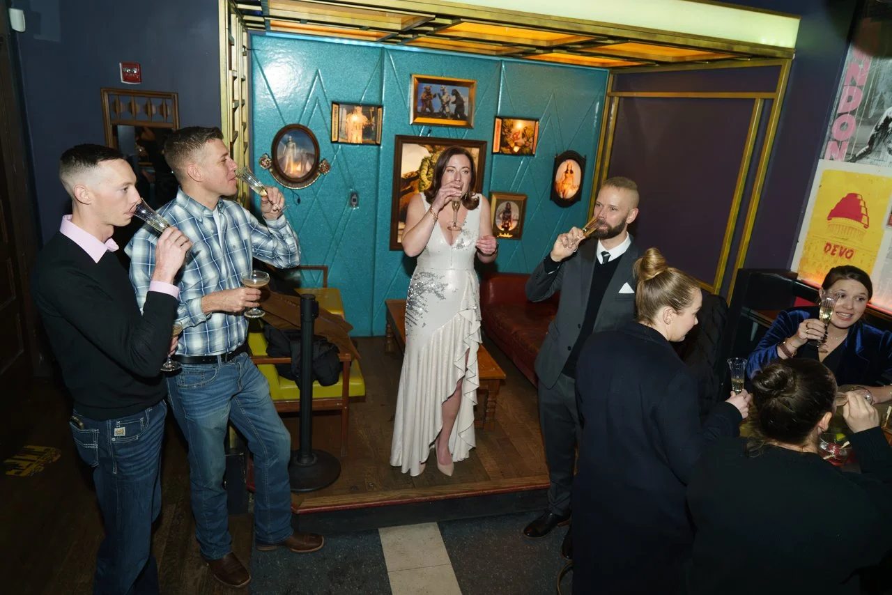 Group of people enjoying drinks at a bar, some standing and some seated, with a woman in a white gown in the center, all sipping champagne in a lively, decorated venue.