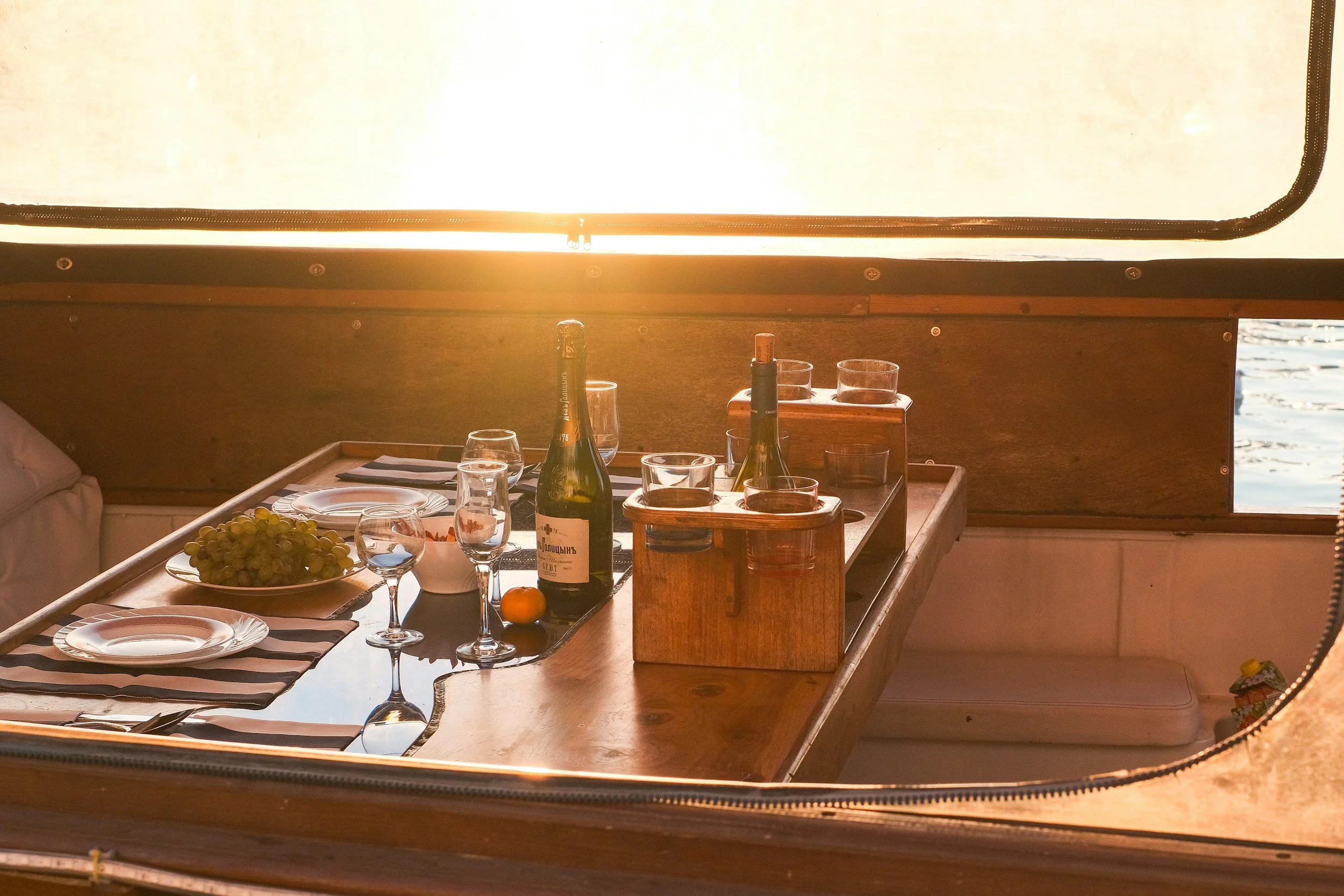 Dinner table with plates, wine glasses, bottles, and a fruit bowl, seen through a boat window with a view of water at sunset.