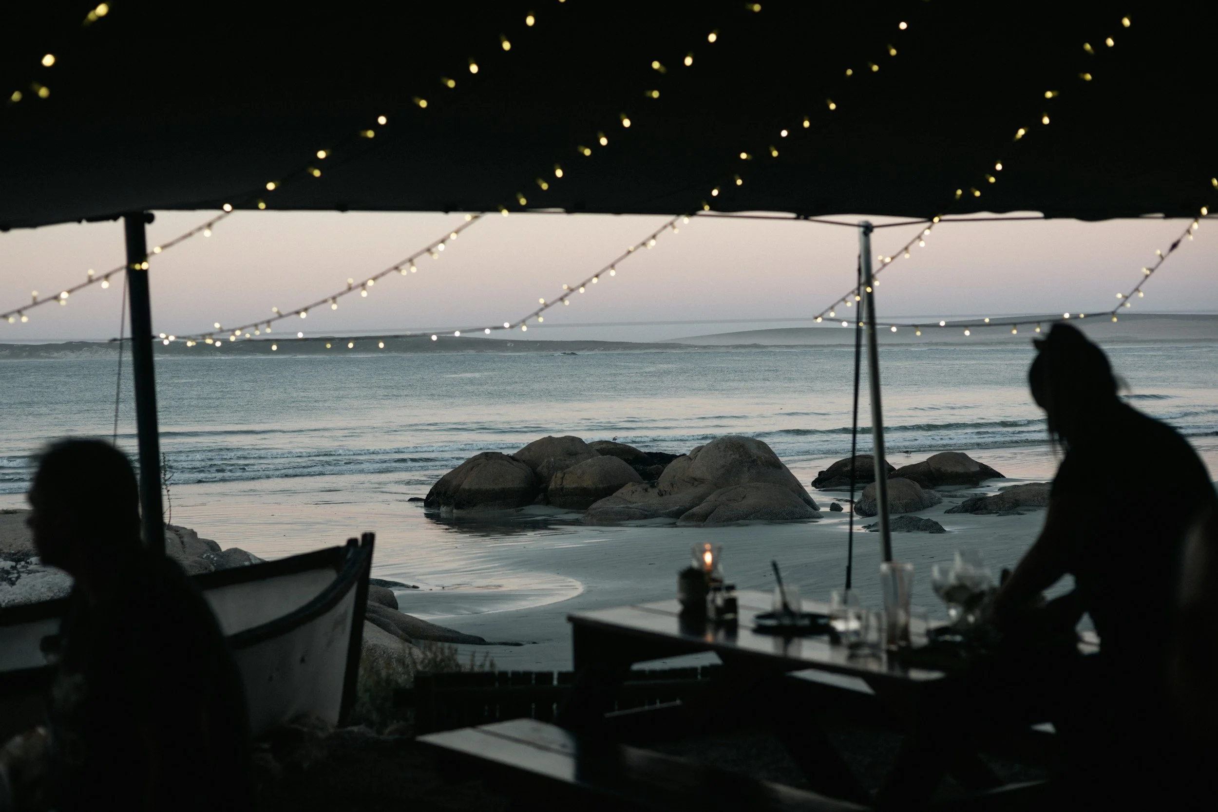 Silhouettes of two people sitting under a canopy with string lights, looking out at the ocean during dusk, rocks visible along the shoreline.