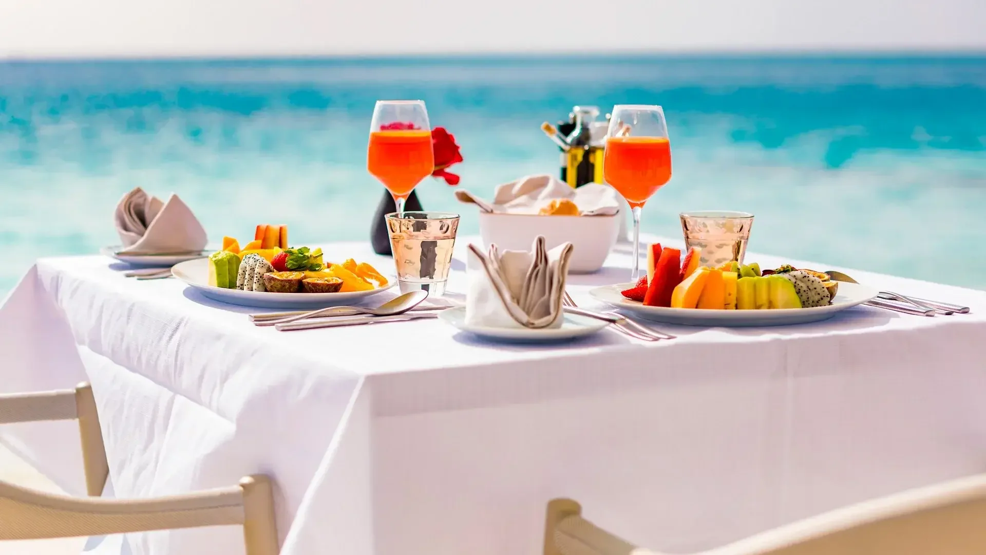 A table set with breakfast food items, including fresh fruit, two glasses of orange juice, water glasses, napkins, and utensils on a white tablecloth, with the ocean in the background.