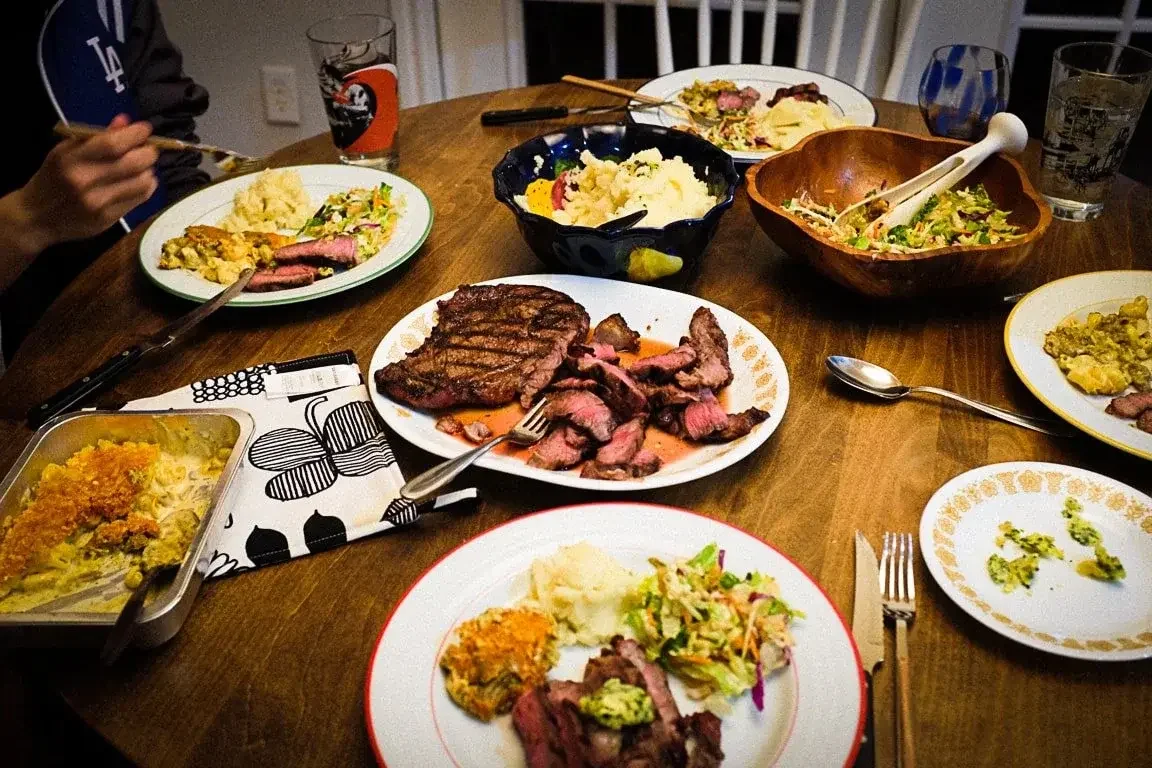 A dinner table with various plates of food including grilled steak, mashed potatoes, salad, and casserole. Two glasses of drinks are also visible.