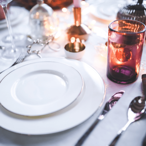 Empty white plate on a table set for dining with a glass, cutlery, and decorative items in the background.