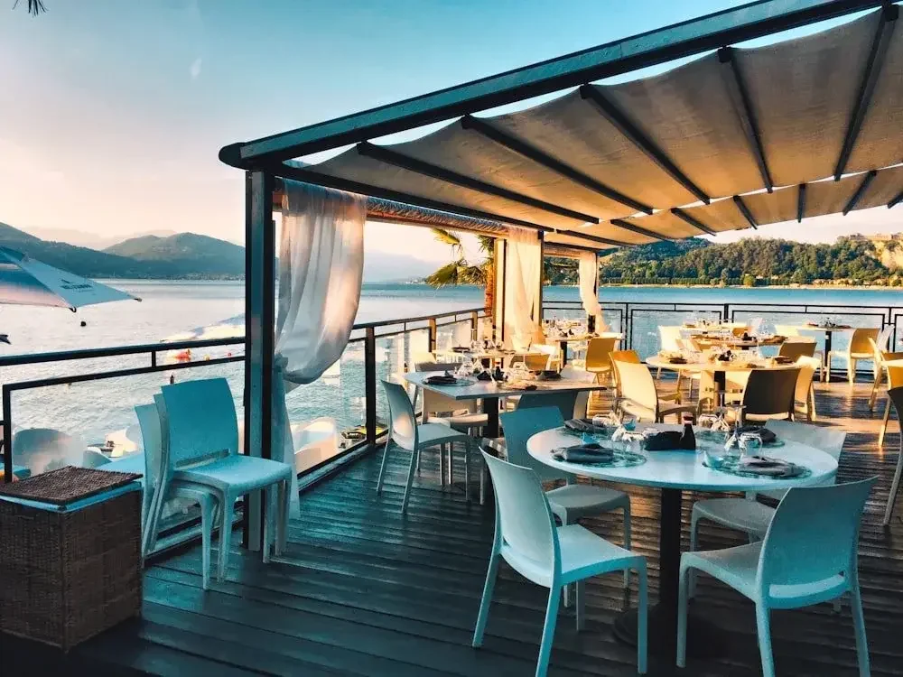 Outdoor restaurant deck overlooking a lake with mountains in the background, set with tables, chairs, and a shaded canopy