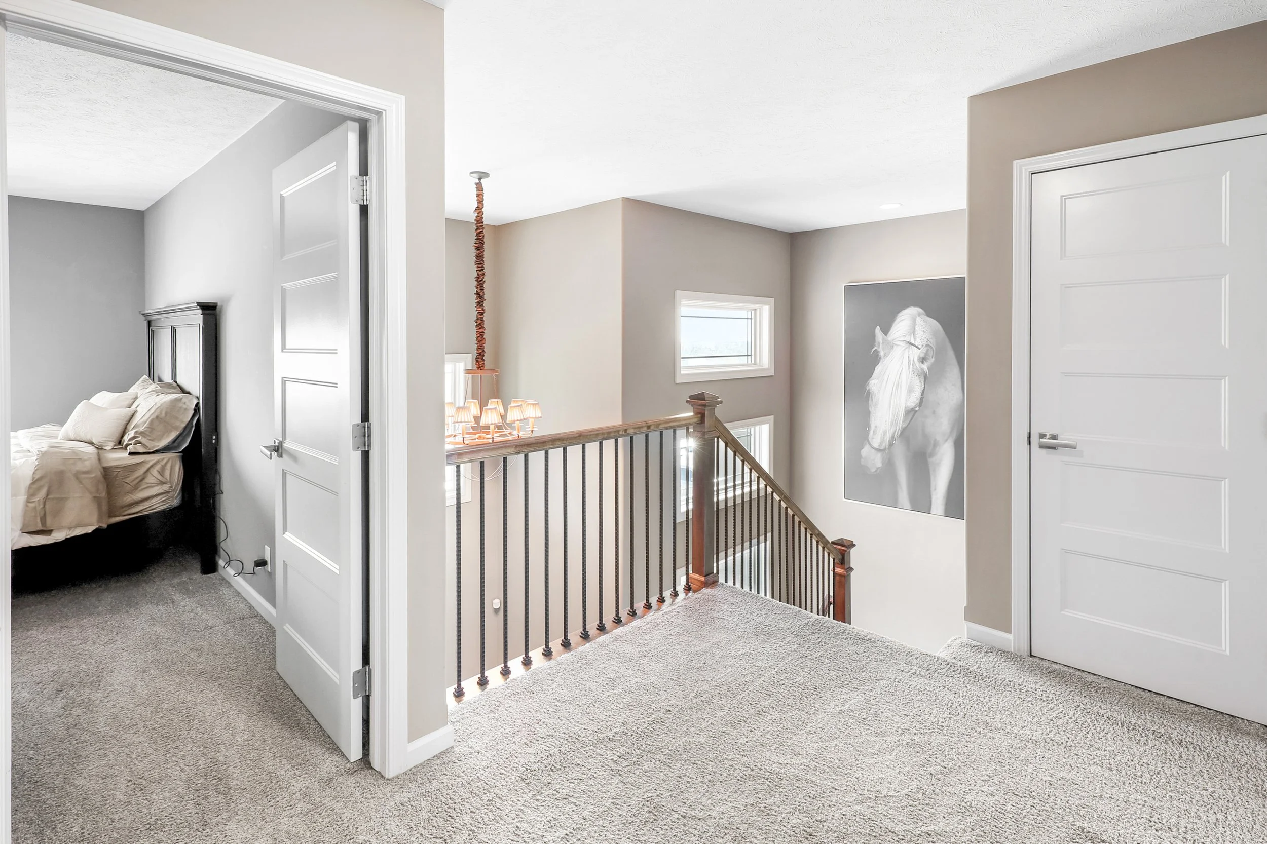 Indoor hallway with beige walls, carpeted flooring, a staircase railing, a framed black-and-white horse photograph on the wall, and an open door leading to a bedroom with a bed.