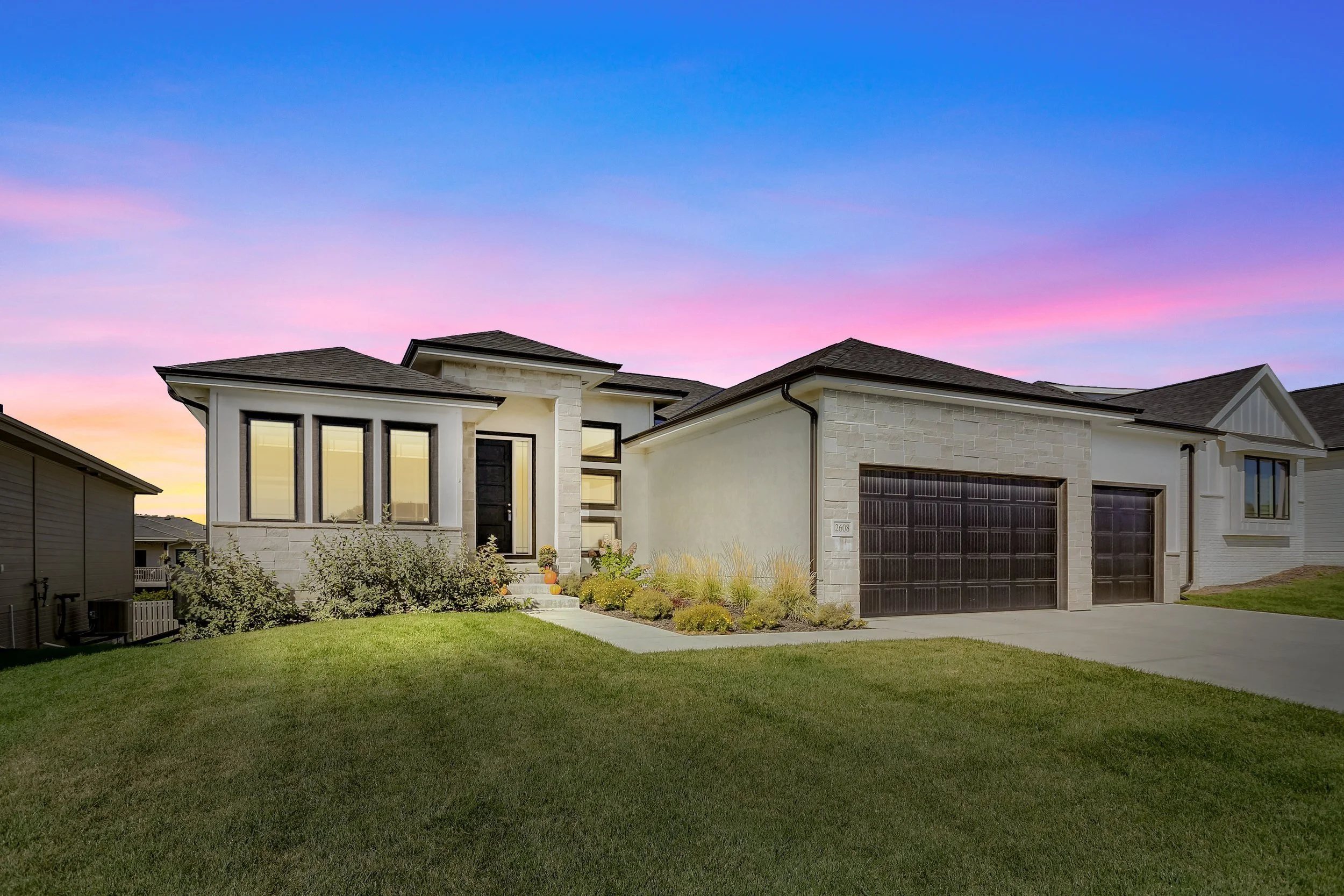Modern single-family home with a manicured lawn, large windows, and a black garage door, set against a colorful sunset sky.