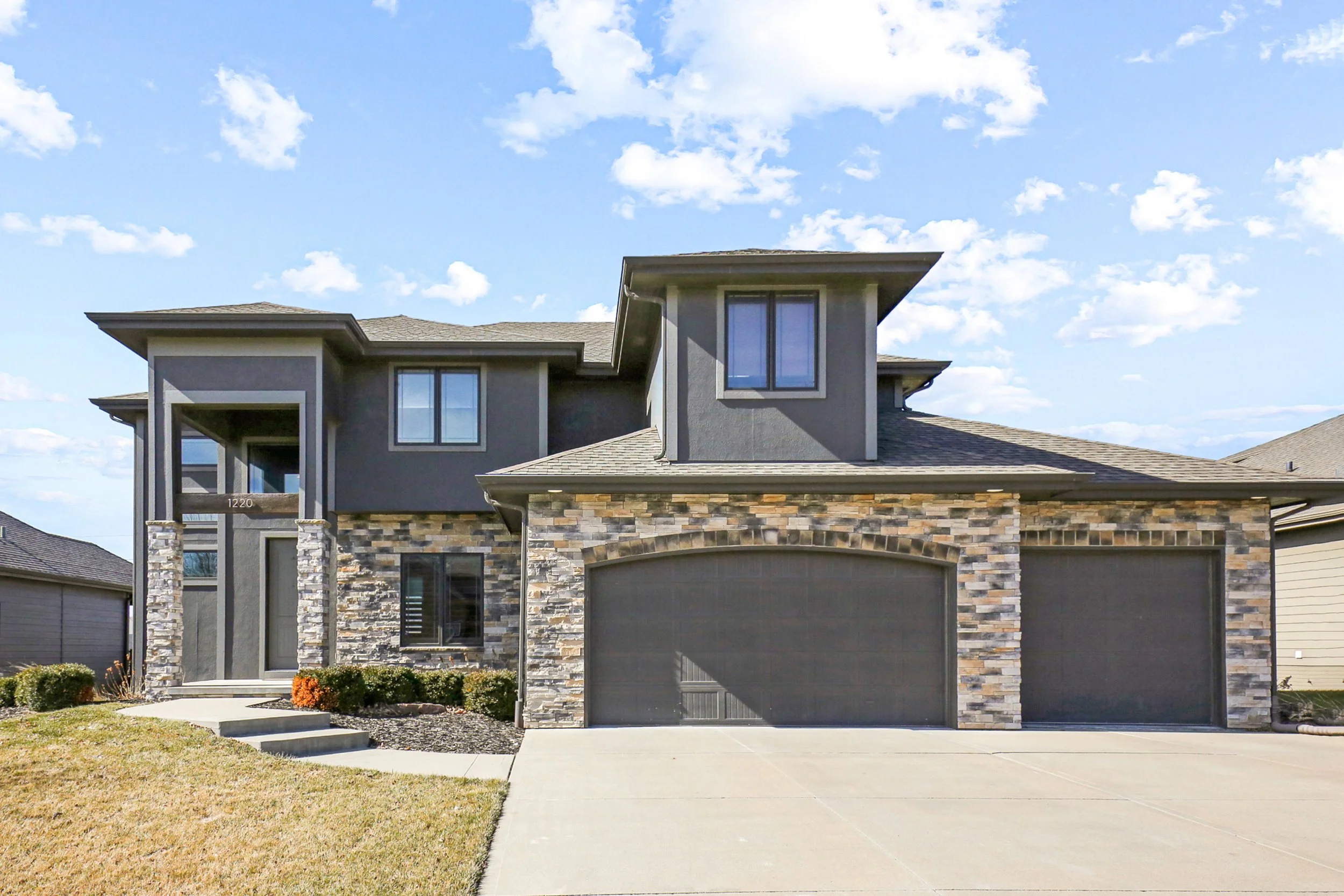 Modern two-story house with a stone facade, dark gray siding, and a three-car garage under a blue sky with scattered clouds.
