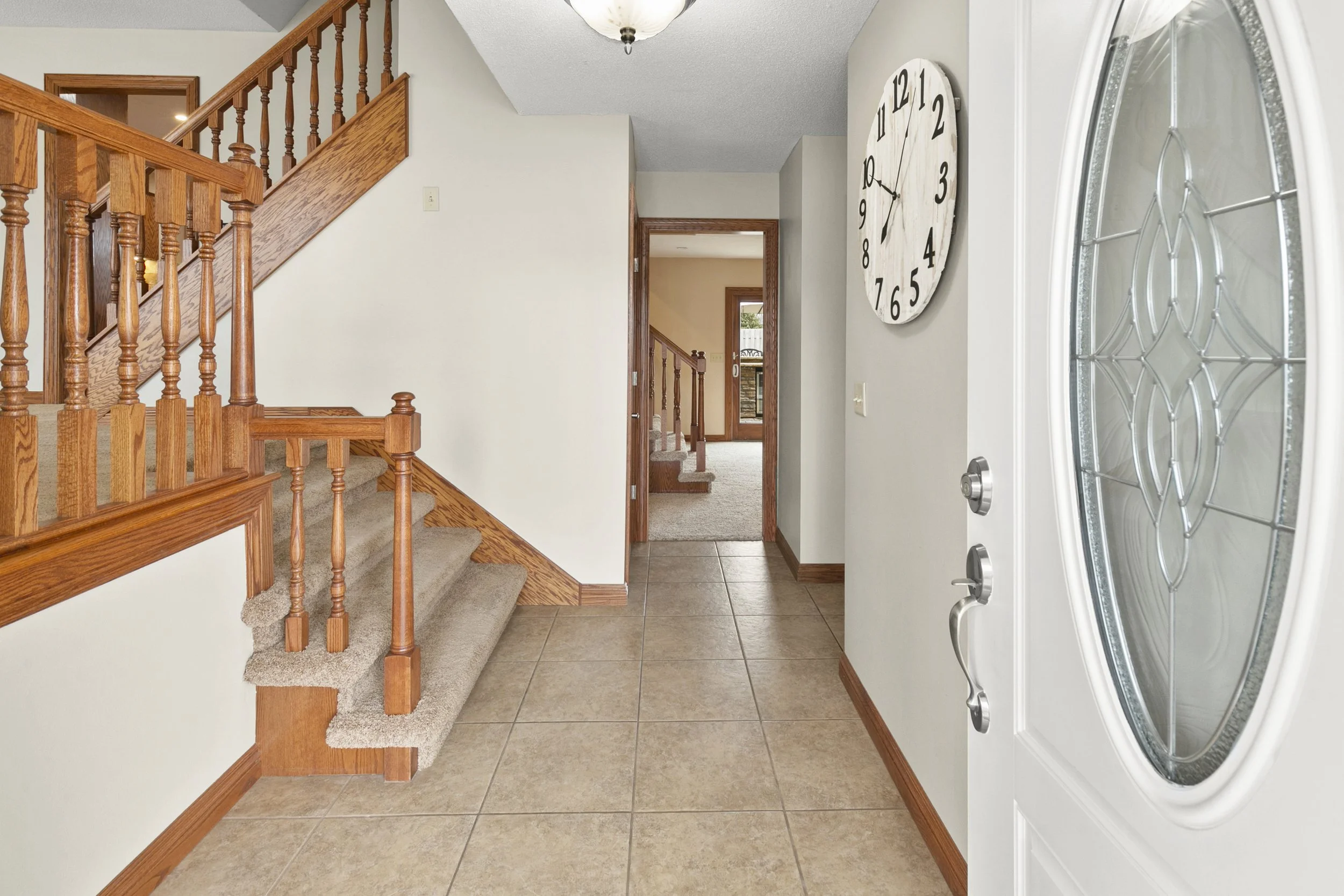 Entryway with a tiled floor, wooden staircase with carpeted steps, a white front door with glass and decorative design, a wall clock, and a view into a living room with a carpeted floor.