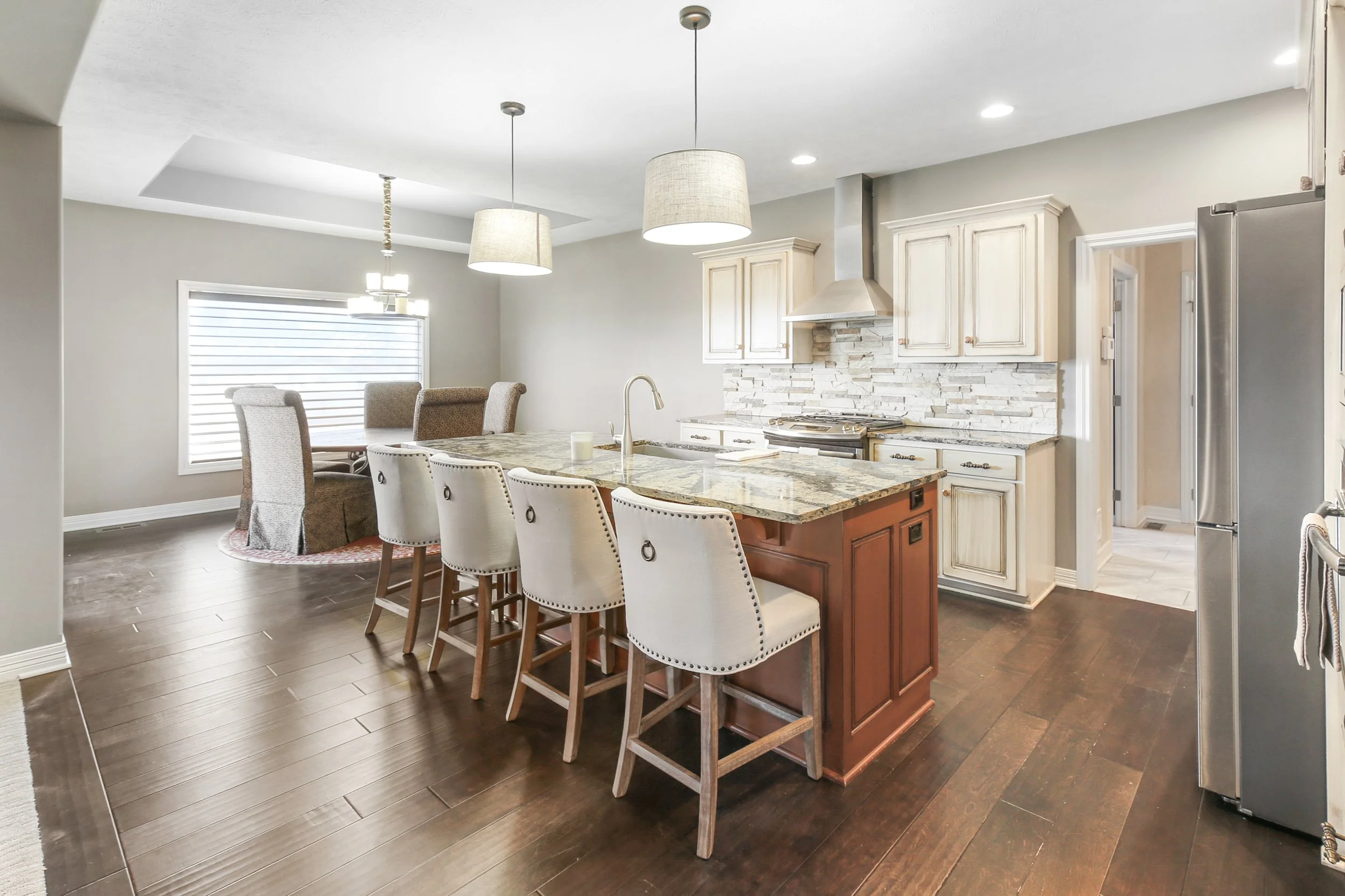 Open-concept kitchen with a large island, white cabinetry, and a stone backsplash. There are six beige upholstered barstools at the island, pendant lights overhead, and a dining area with a circular table and six chairs near a window with blinds. Dark wood flooring runs throughout.