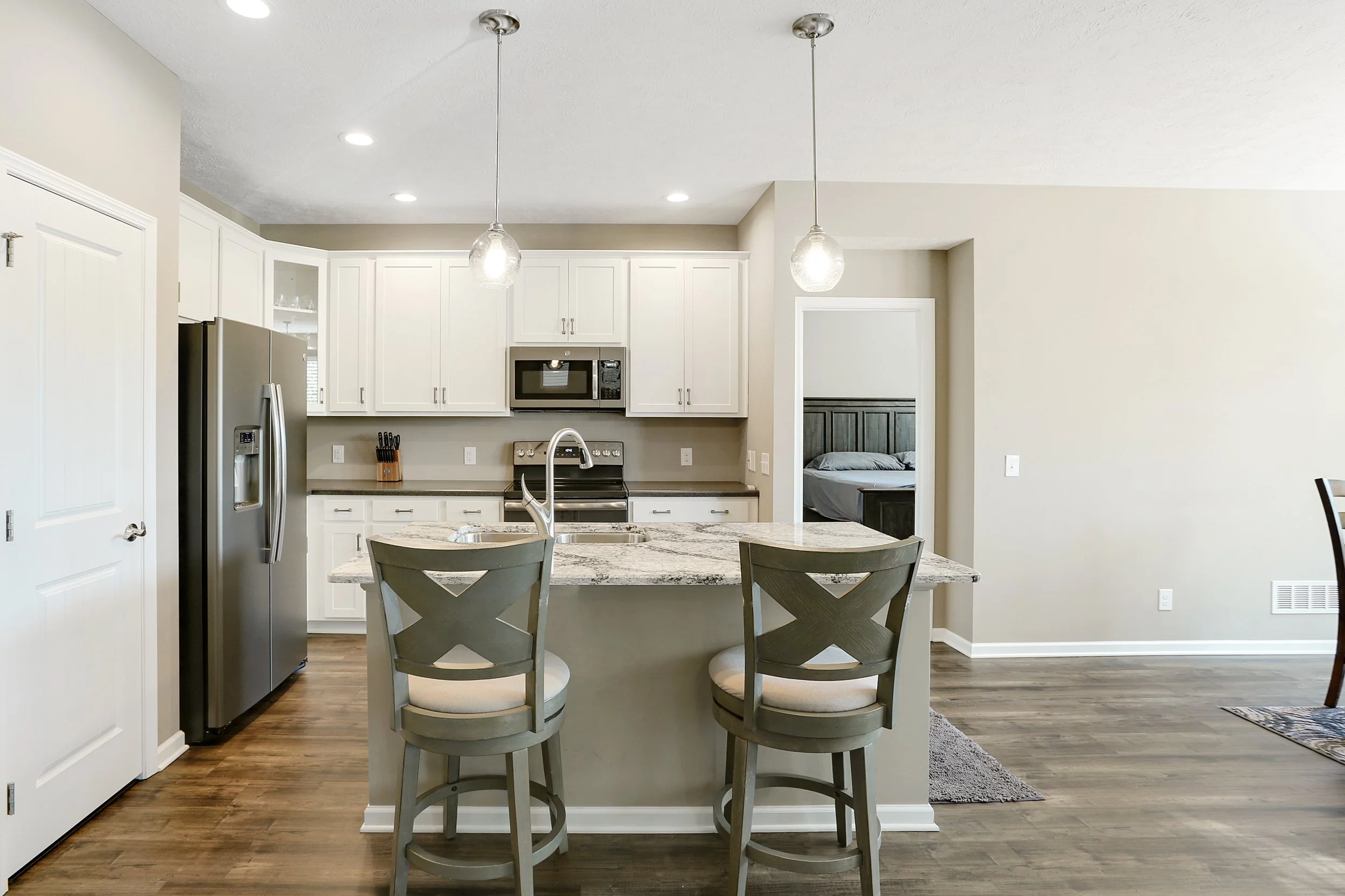 Modern kitchen with white cabinets, stainless steel refrigerator, black appliances, granite counter island, two barstools, and a view into a bedroom with a dark wood headboard.