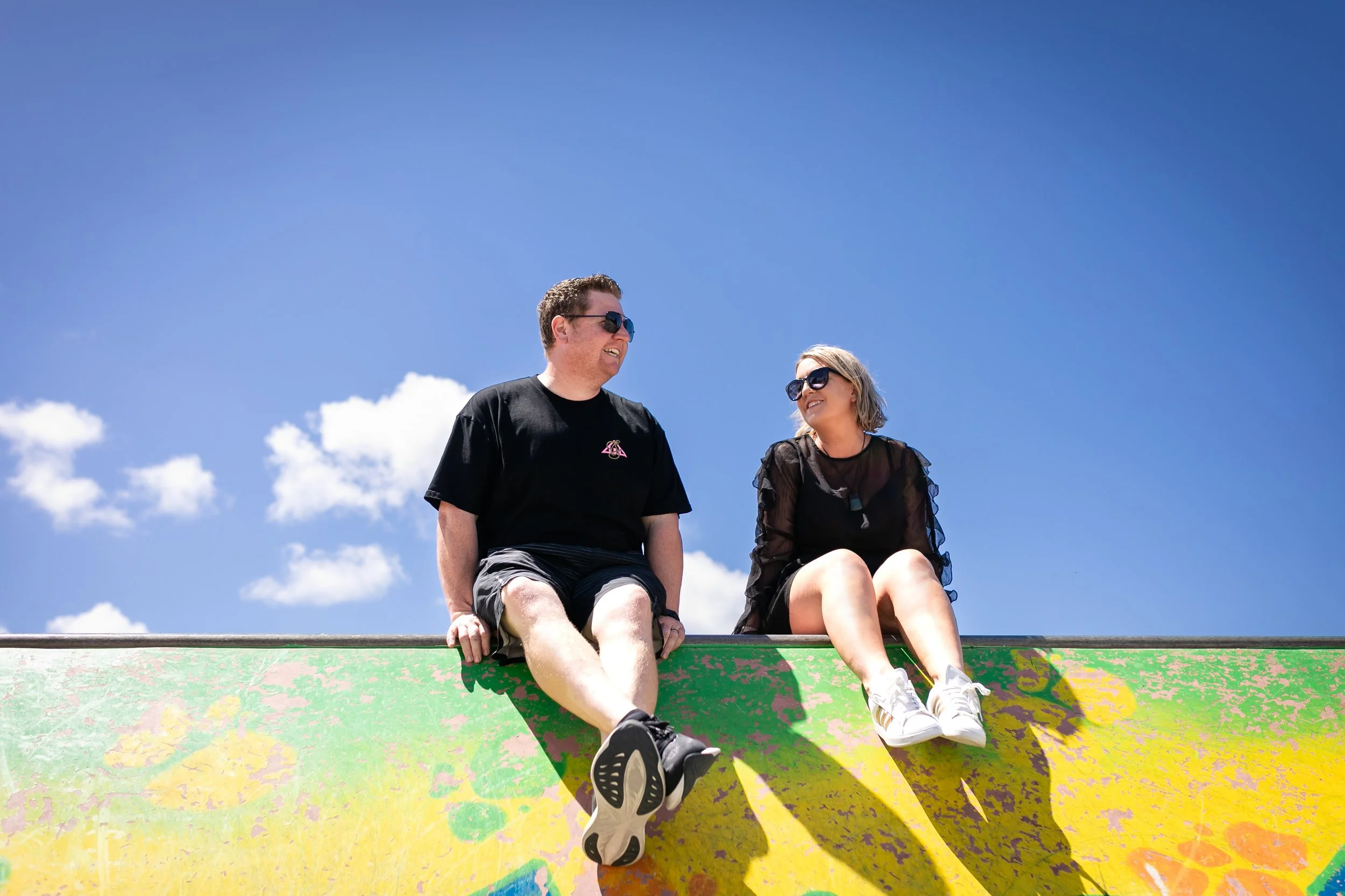 A man and woman sitting on the edge of a colorful skate ramp, smiling and talking, under a clear blue sky with a few clouds, wearing sunglasses and casual clothes.