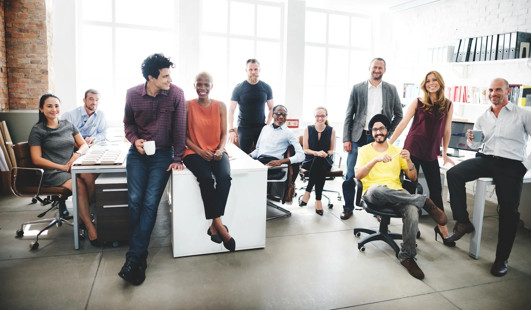 A diverse group of ten people in an office, some sitting and some standing, smiling and gathered around a desk in a bright room with large windows and white walls.
