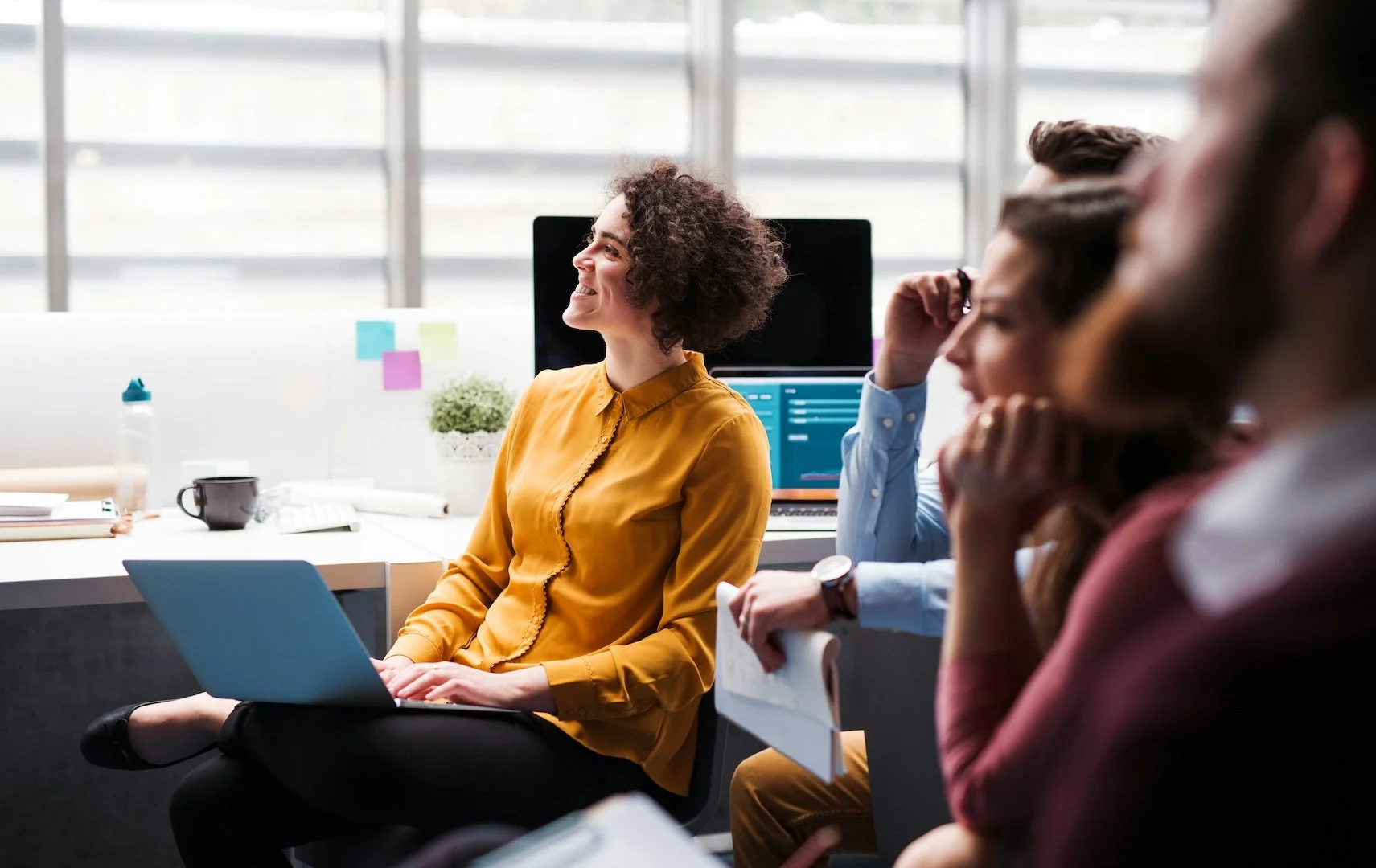 Group of people in a business meeting or workshop, with a woman in a yellow blouse smiling and sitting with a laptop, others listening attentively in a modern office setting.