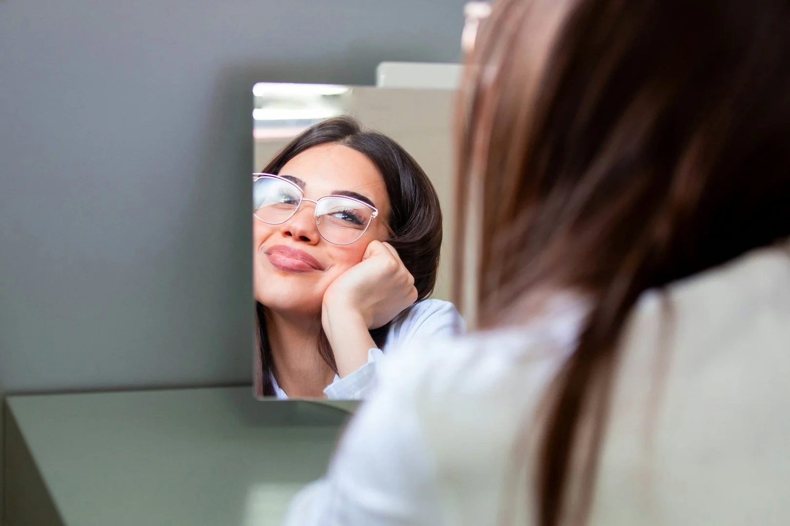 A woman with glasses resting her face on her hand, looking at her reflection in a mirror.