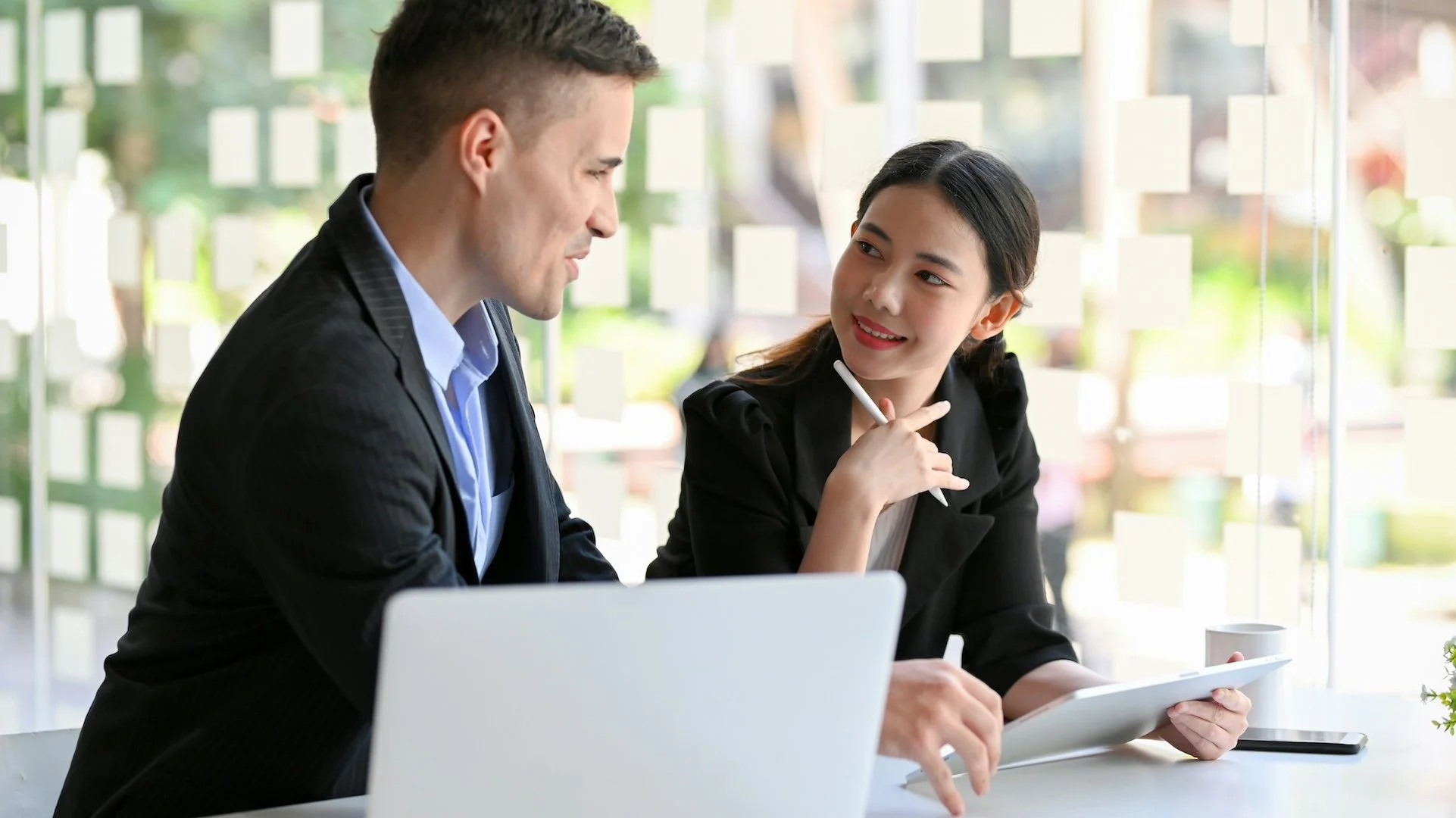 Two business professionals, a man and a woman, having a discussion at a table in a bright office. The woman is smiling and holding a tablet, while the man is pointing at it with a pen.