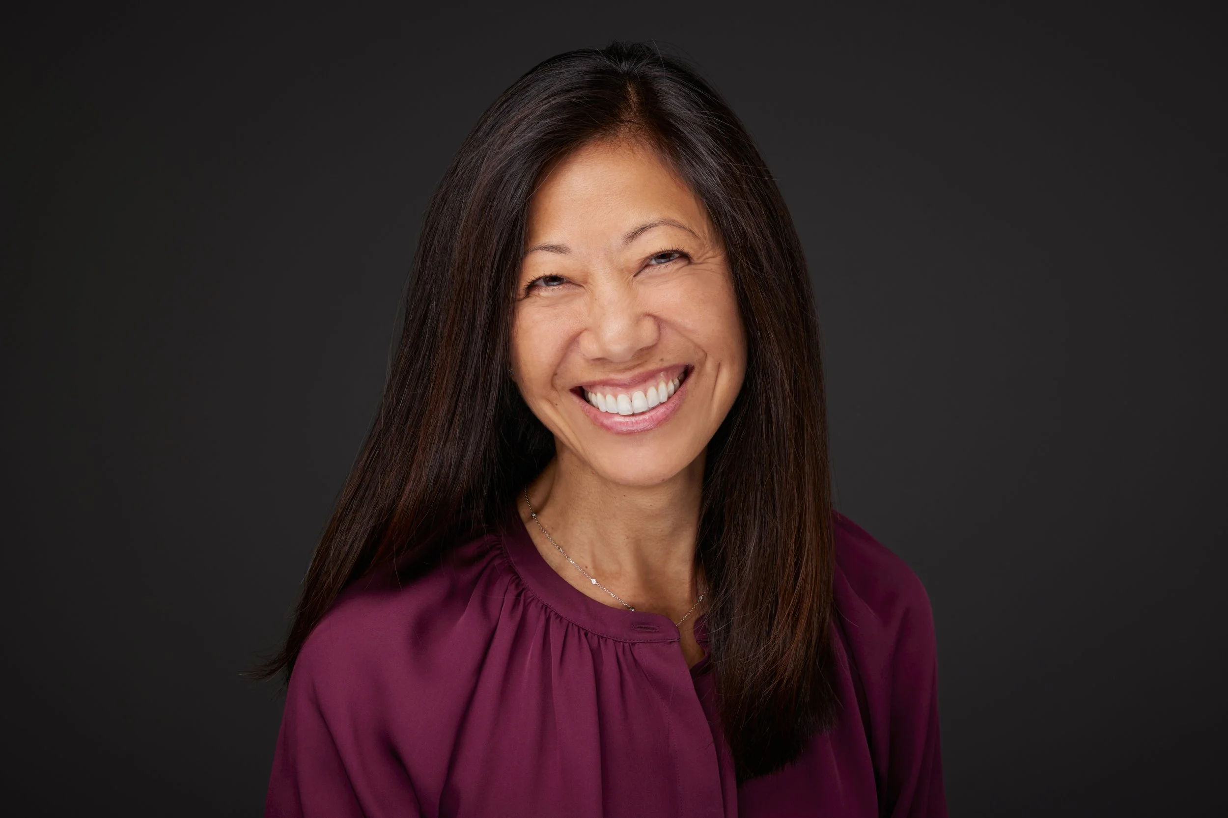 A woman with long dark hair smiling against a dark gray background, wearing a maroon blouse.