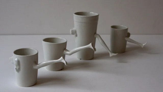 Ceramic cups connected by Barbie doll legs, arranged in a line on a neutral surface, transforming domestic vessels into fragile, body-like forms.