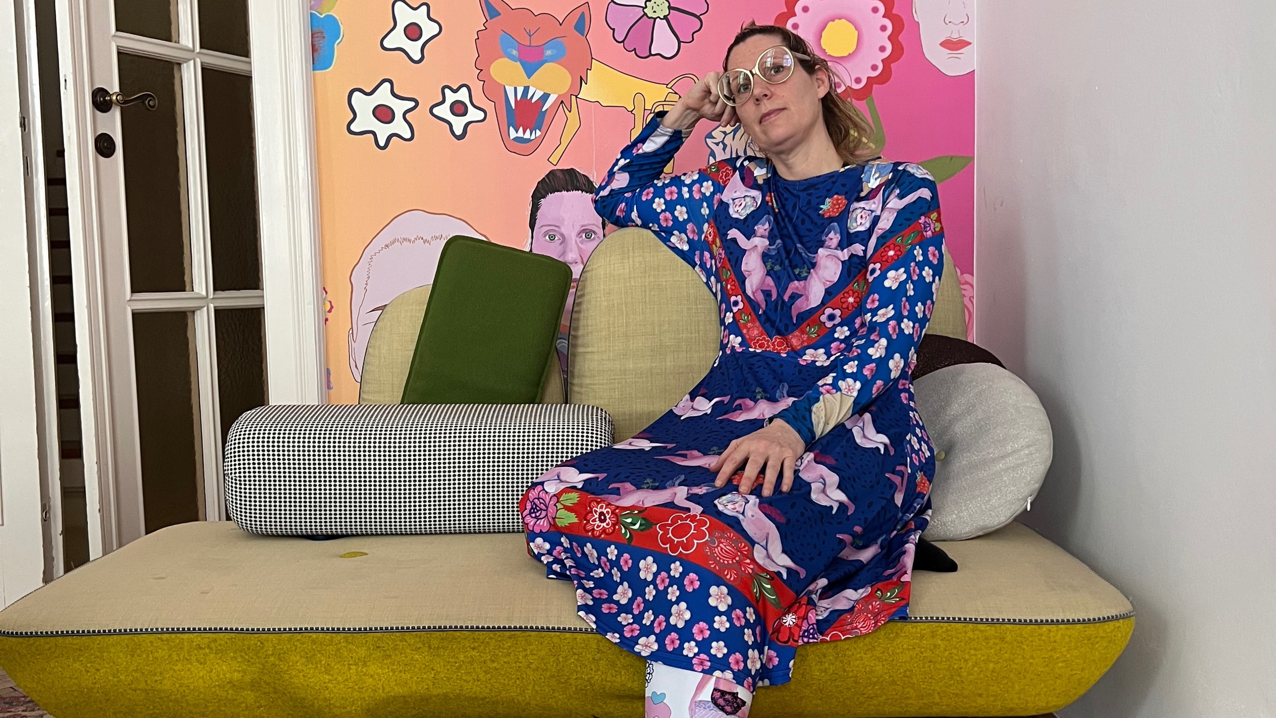 Eine Frau in einem bunten, gemusterten Kleid sitzt auf einem gelben Sofa vor einer pinken Wand mit bunten, abstrakten Kunstwerken und Tiermotiven.