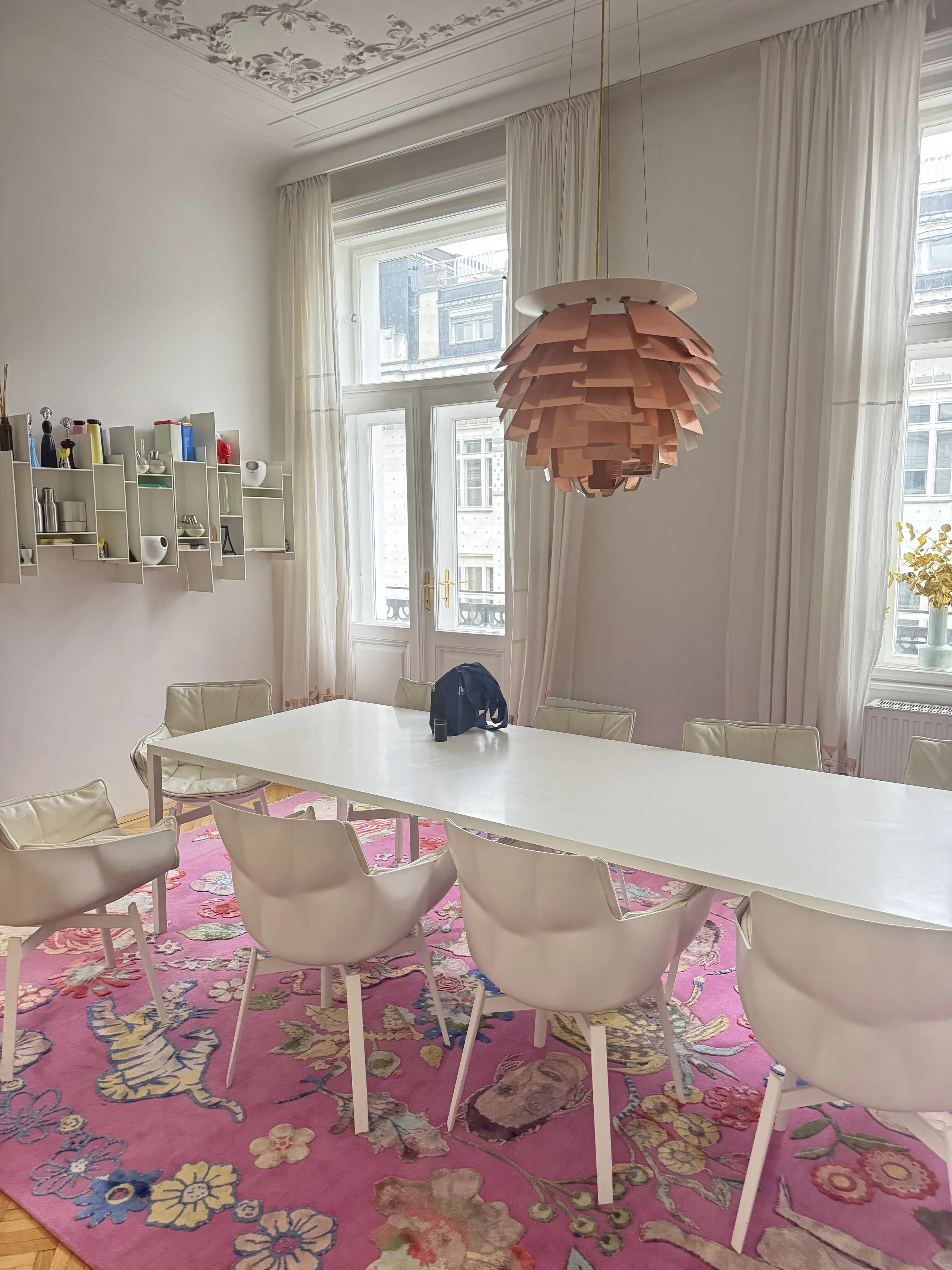 Ein heller Esszimmer mit großen Fenstern, weißen Wänden und einer rosa, mit Blumen bedruckten Teppich. Ein langer weißer Tisch ist von weißen Stühlen umgeben, und eine moderne hölzerne Deckenlampe hängt darüber. Es gibt eine weiße Wandregal mit Dekorationsgegenständen an der linken Wand.