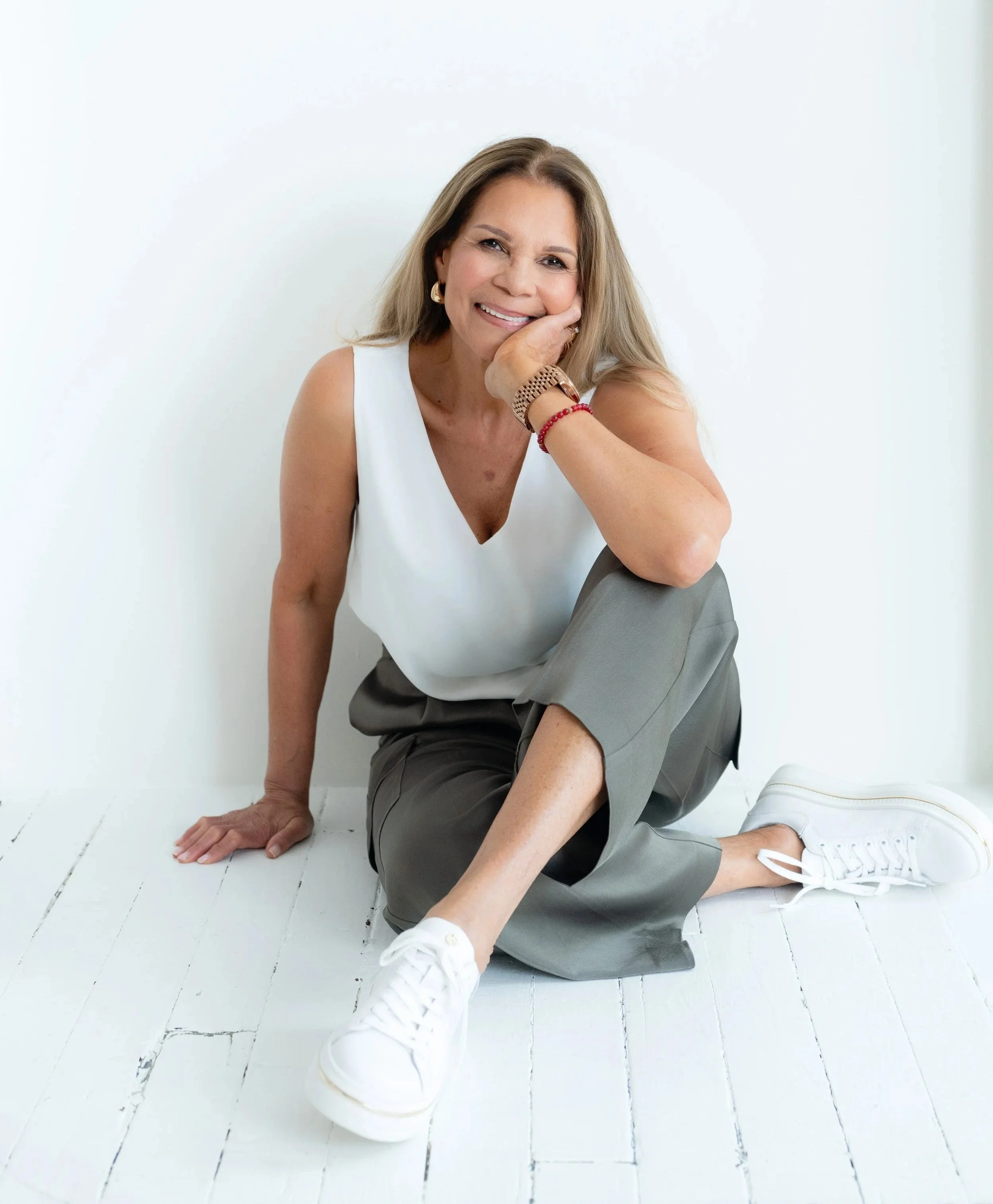 A woman with blonde hair, wearing a white sleeveless top, gray pants, and white sneakers, sitting on a white wooden floor with her head resting on her hand, smiling at the camera against a plain white background.